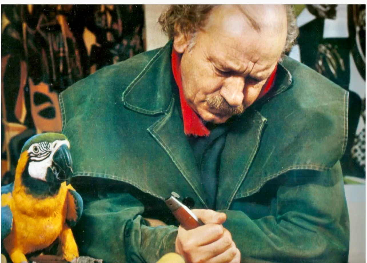 A man with a mustache and curly hair, wearing a green jacket, holds a pen and is looking down. A colorful parrot is perched on his arm, close to his hand. The background has various objects, possibly in a store or workshop.
