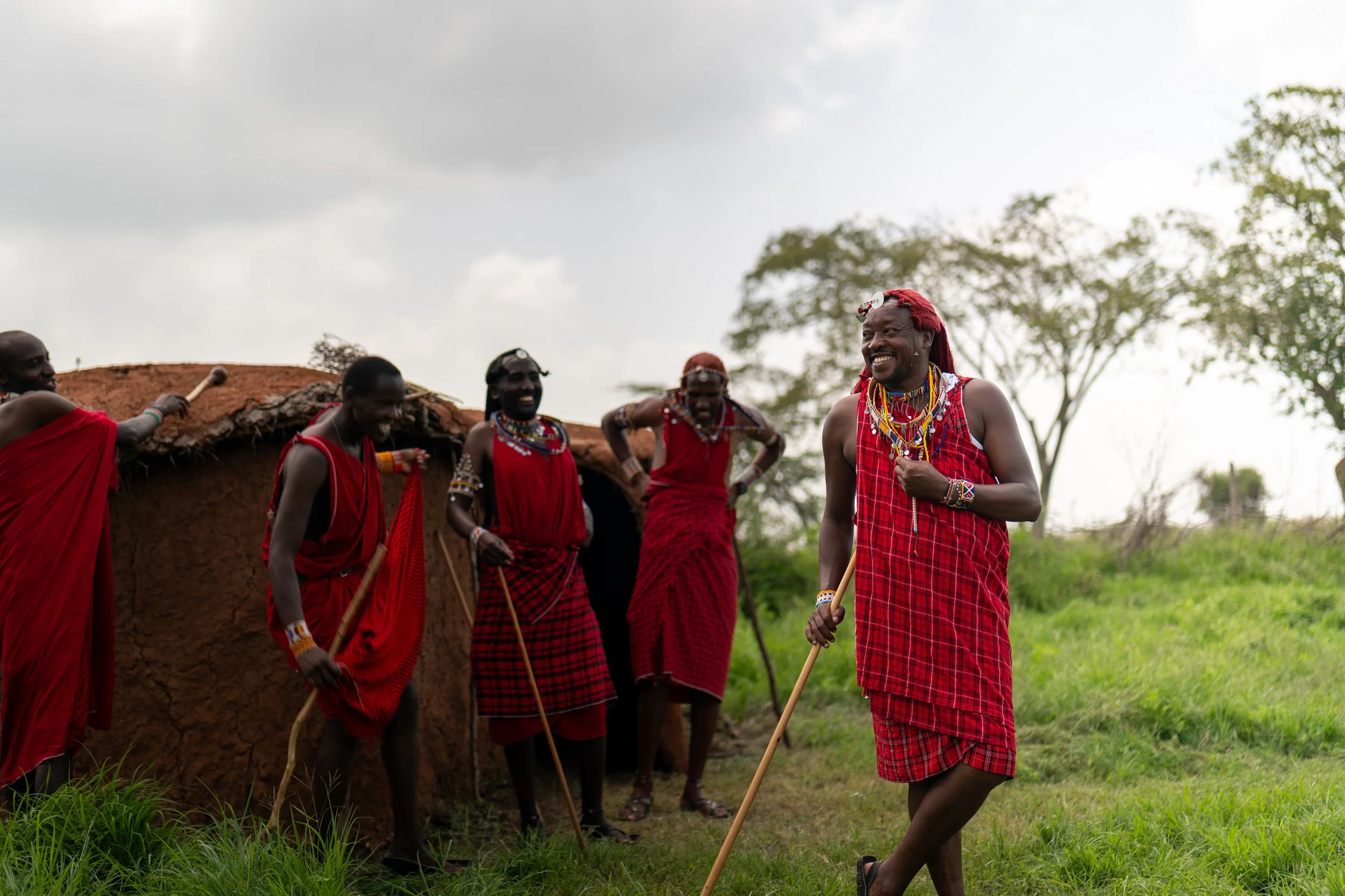 Group of Maasai men wearing red shuka robes and traditional jewelry, standing outdoors on green grass with trees and cloudy sky in the background, engaging in a cultural activity.
