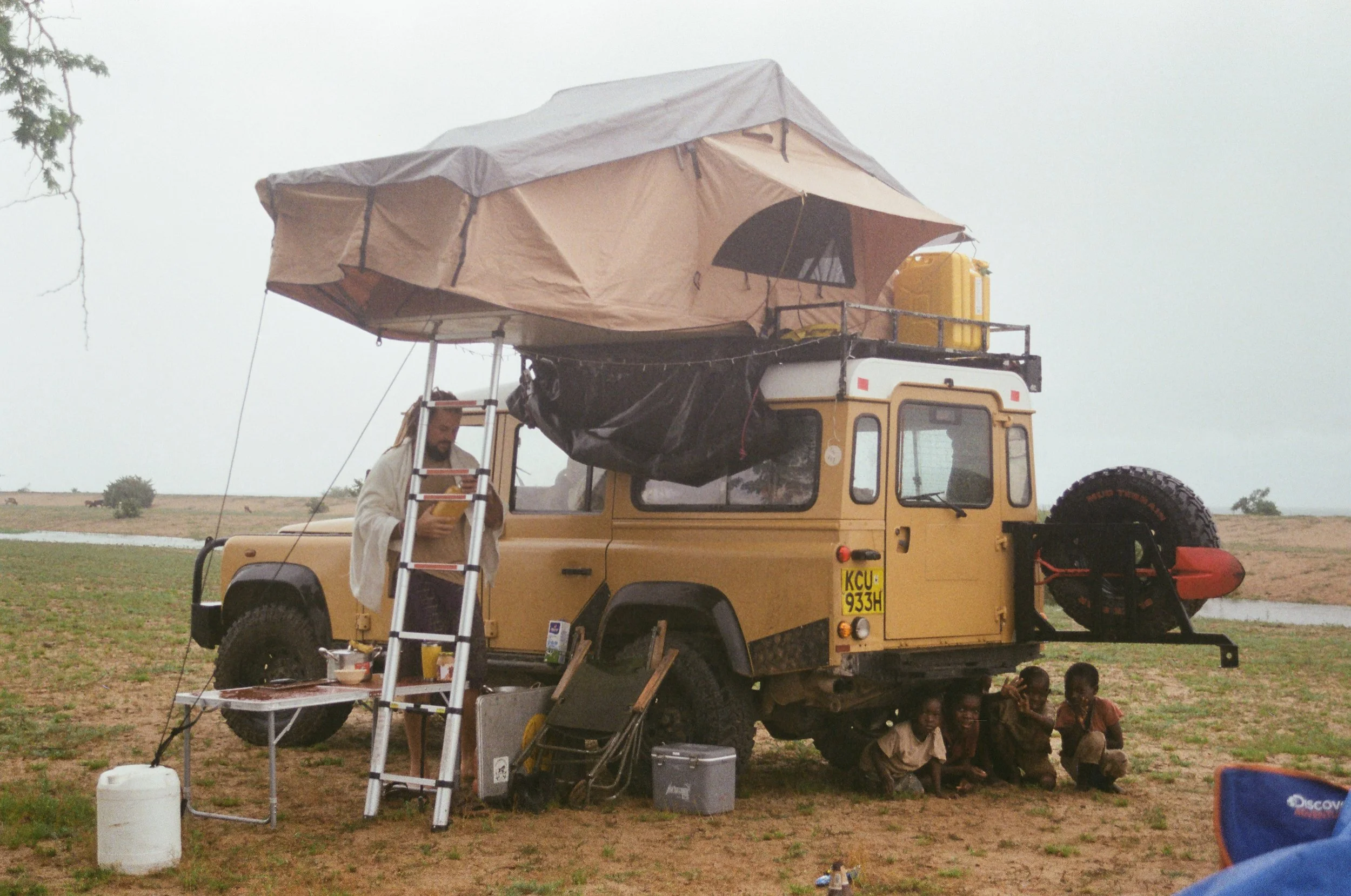 A yellow off-road vehicle parked on a grassy field with a rooftop tent, ladder, and camping equipment underneath. Several children are sitting on the ground near the vehicle.