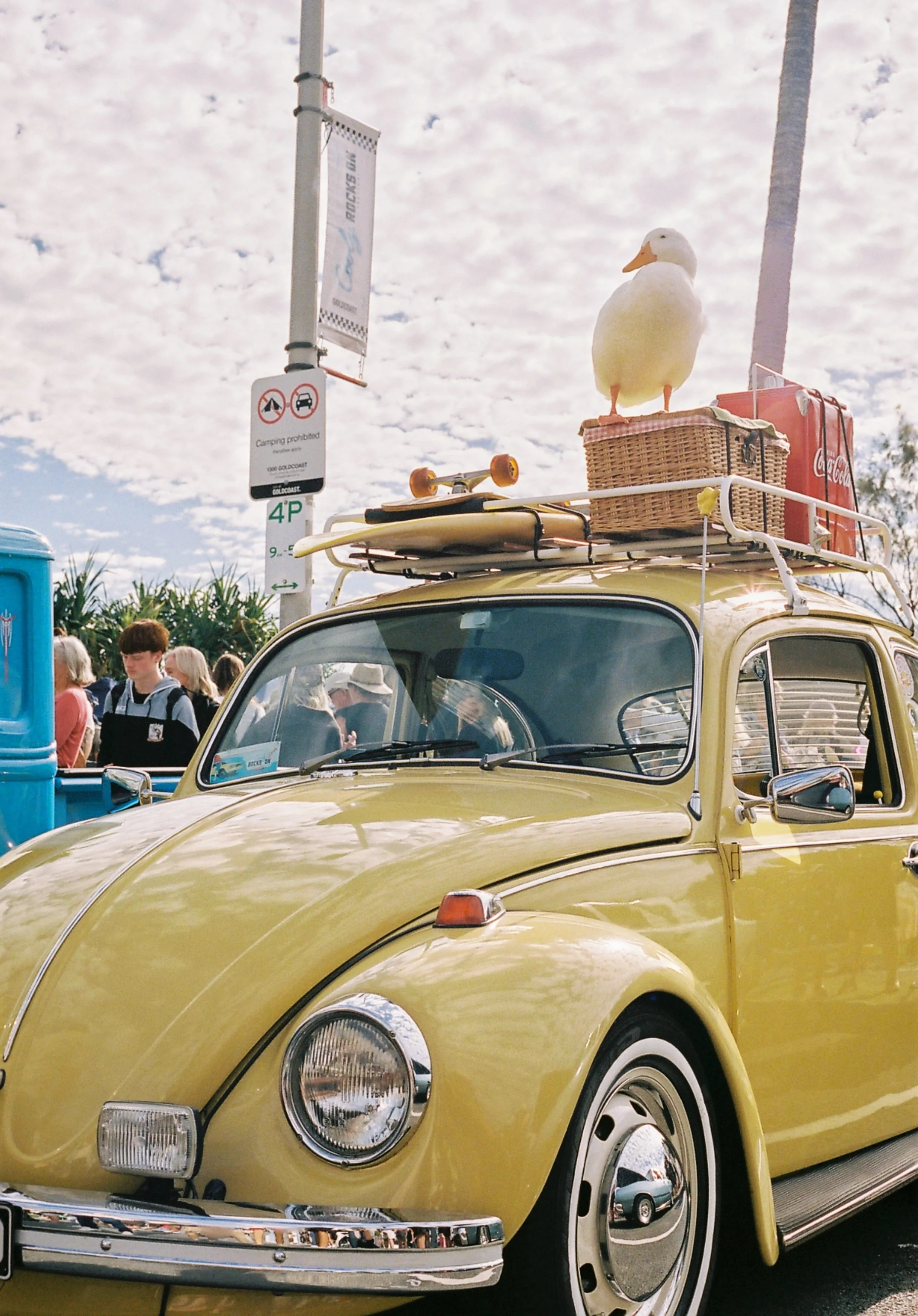 Close-up of a vintage yellow Volkswagen Beetle car with a round headlight and chrome bumper, parked outdoors during daytime. In the background, there are people and a street sign with a partly cloudy sky.