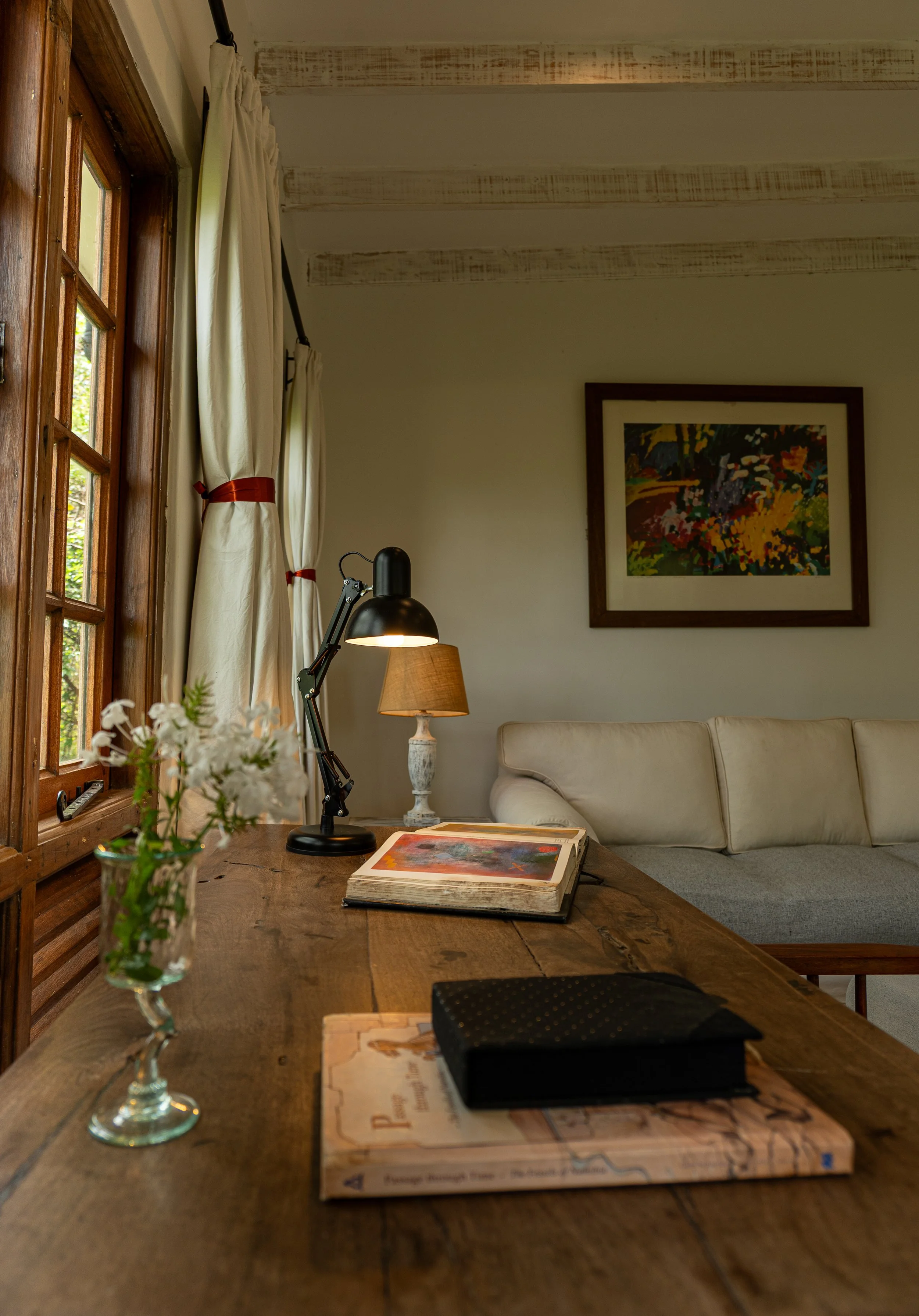 Interior of a cozy room with a wooden desk, a potted plant, a closed book, and a black box, with a window, cream-colored curtains, a floor lamp, and a painting on the wall.