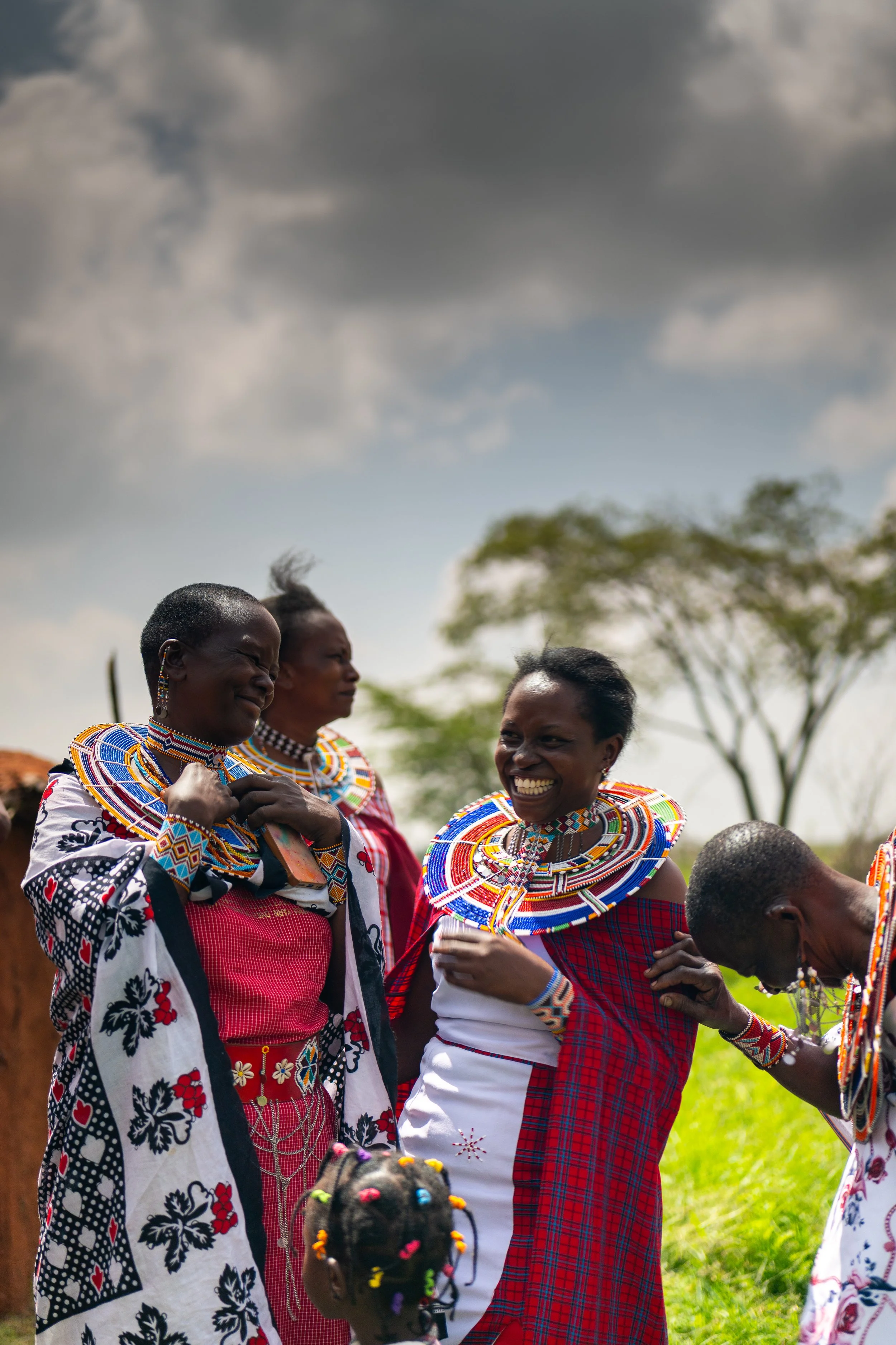 A group of women in colorful traditional Maasai clothing, smiling and engaging in a cultural gathering outdoors under a cloudy sky, with greenery and a tree in the background.