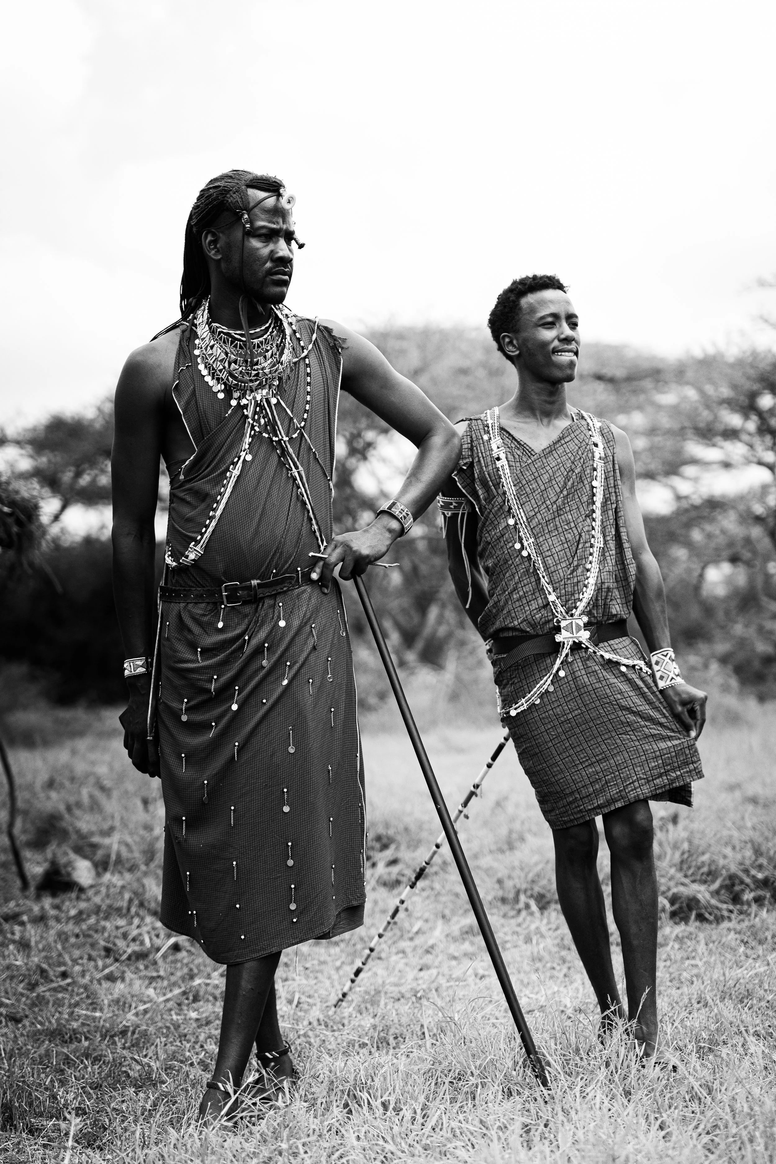 Two Maasai men dressed in traditional attire standing outdoors in a grassy area, holding a stick, with trees in the background.