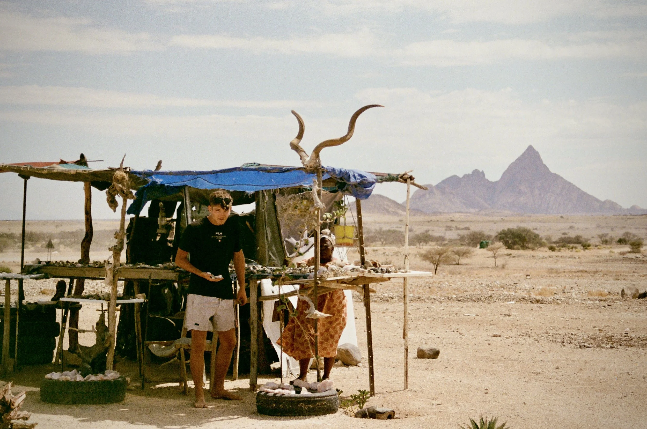 A small roadside market stall in a desert landscape with a mountain in the background, displaying various items for sale, and two children standing nearby.