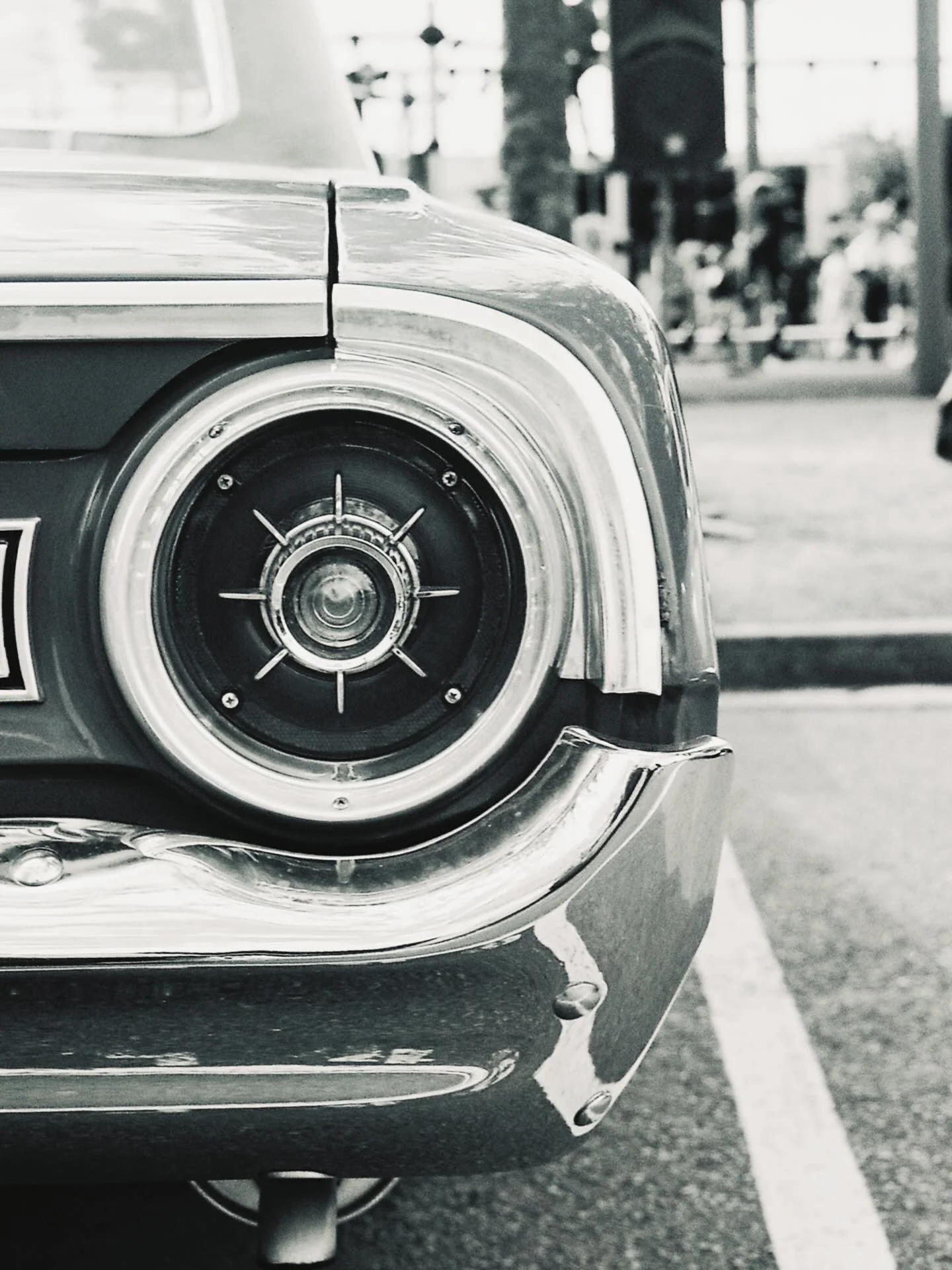 Close-up of the front left corner of a vintage car, focusing on the headlight and front bumper, parked in a lot with other cars and people in the background.