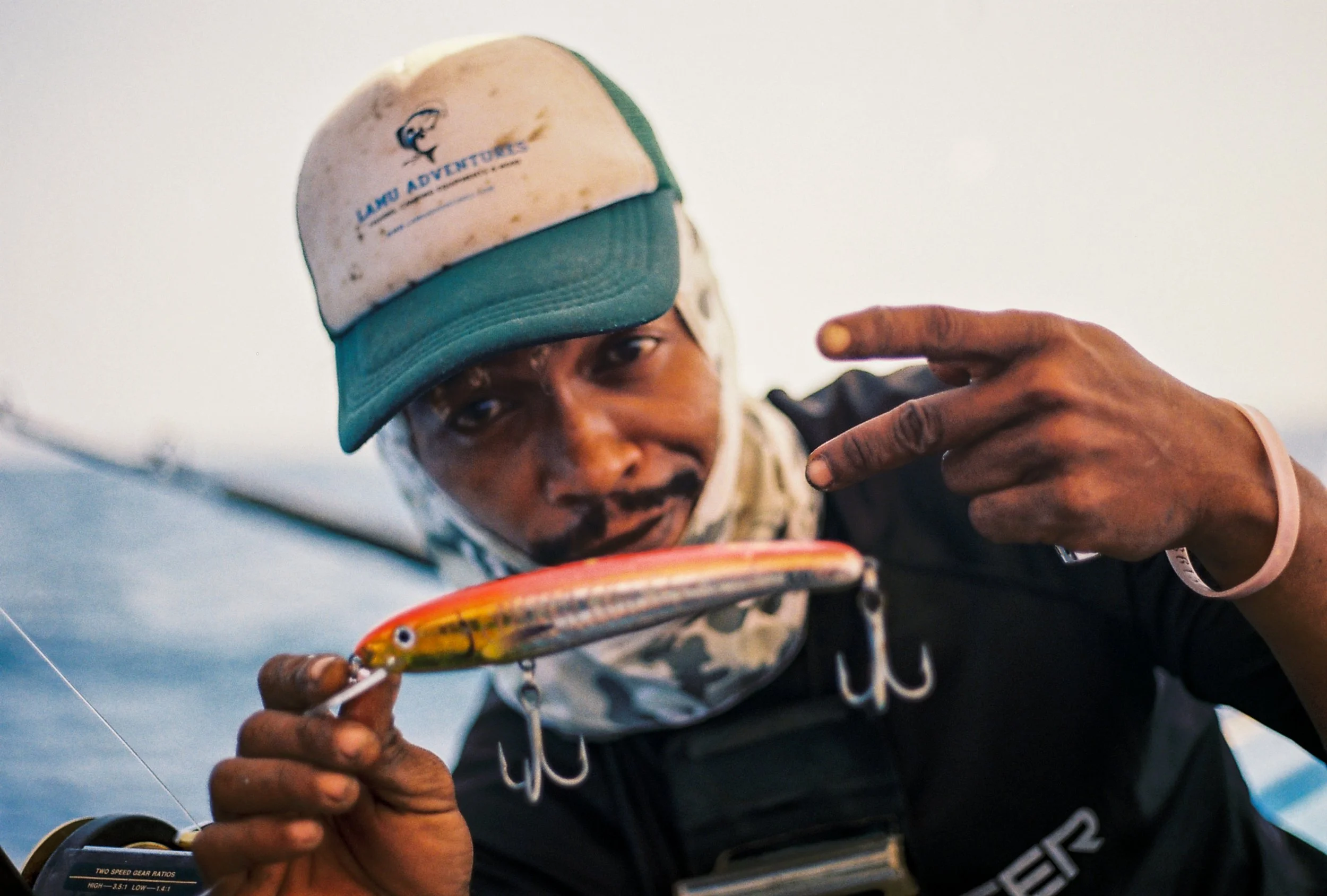 Man wearing a baseball cap with 'LANO Adventures' logo, holding a colorful fishing lure, with a sky background.