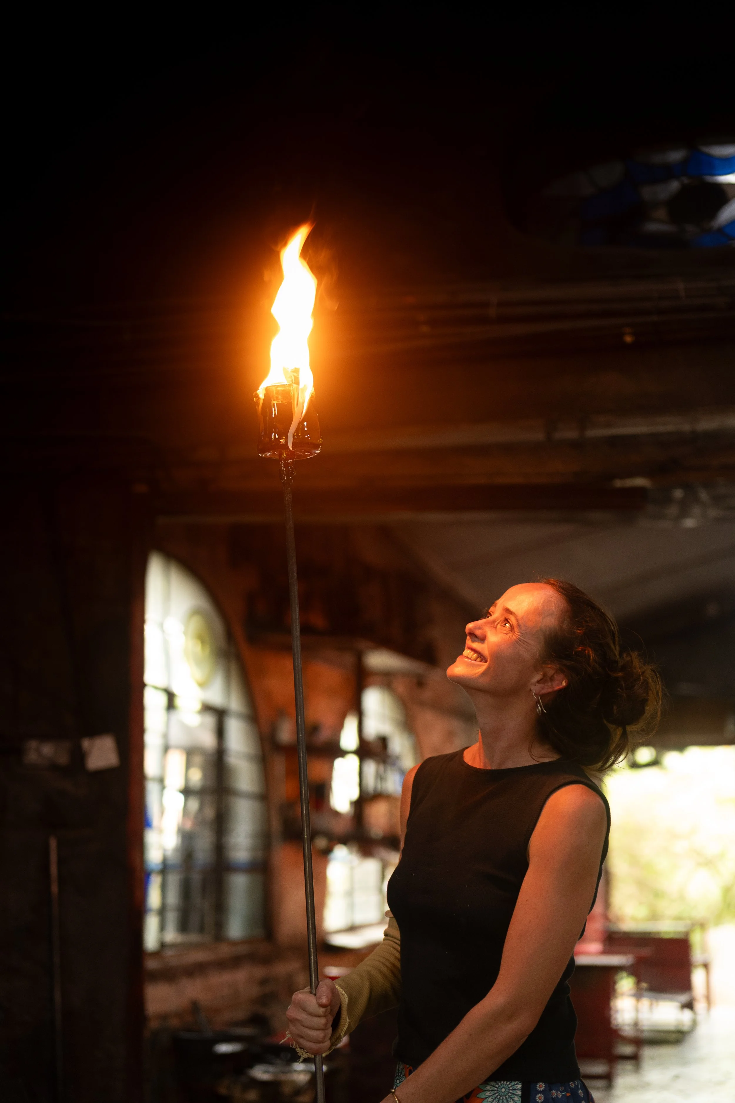 A woman smiling and looking up while holding a lit torch with a bright flame indoors.