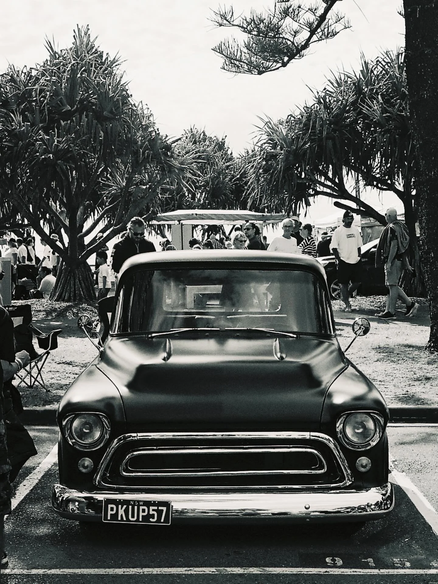 Black and white photo of a vintage car parked in front of a busy outdoor market with people walking among trees and stalls.