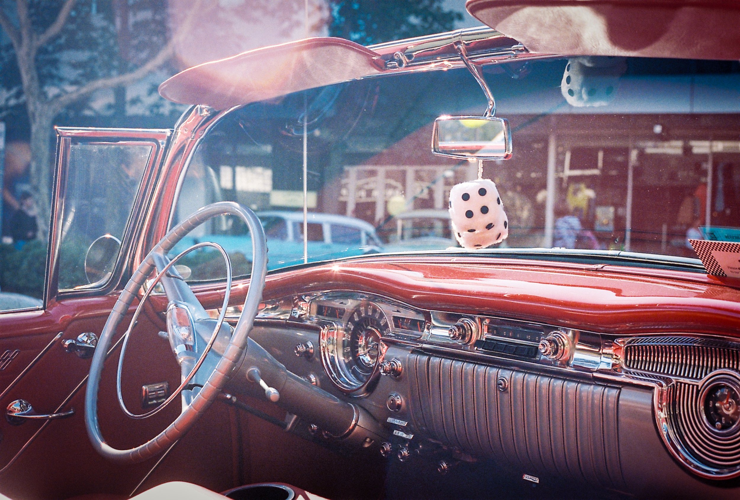 Interior of a vintage red car with a white fuzzy dice hanging from the rearview mirror, car dashboards, and steering wheel.