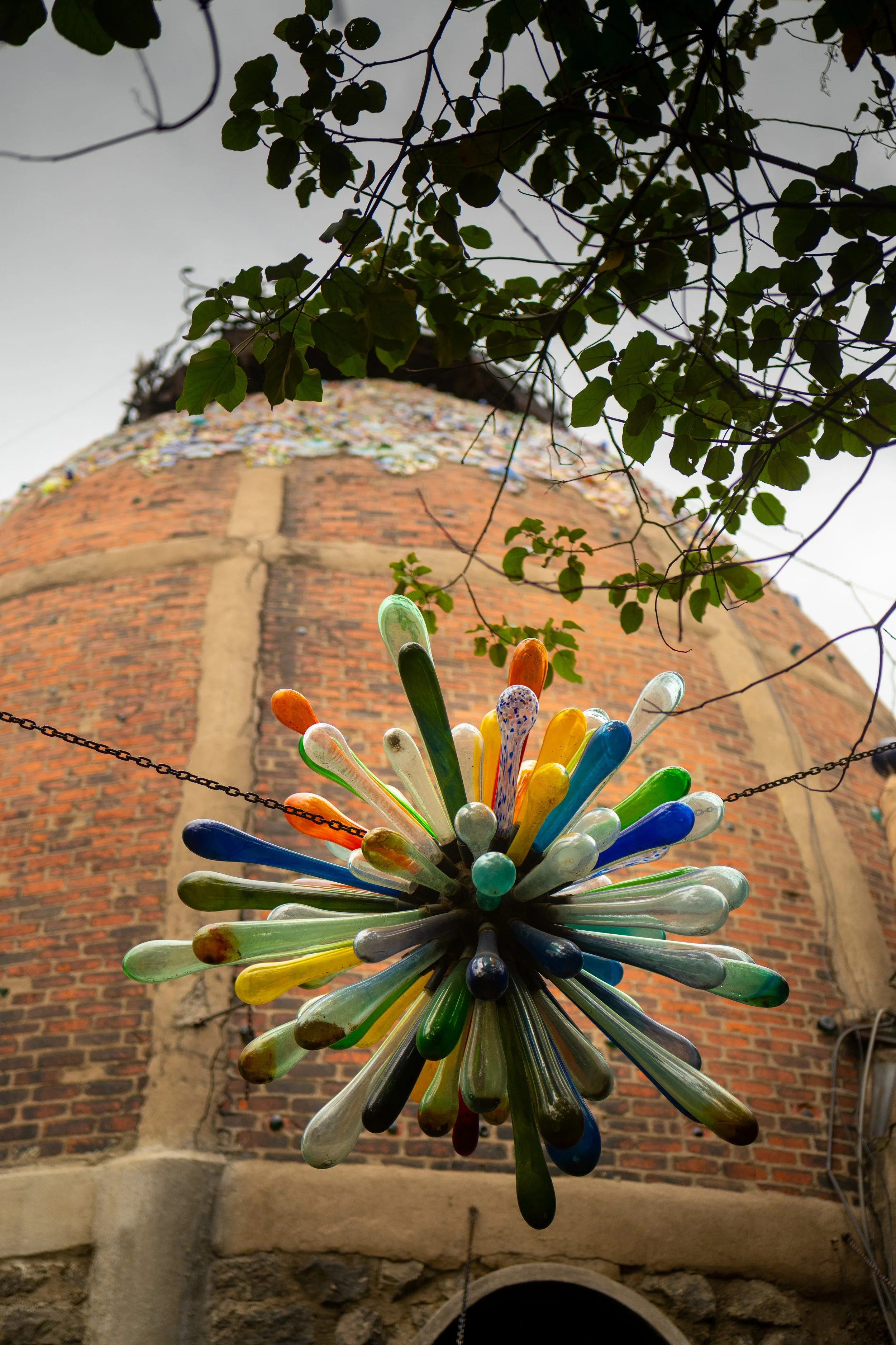 Colorful glass art sculpture hanging in front of a brick and stone tower, view from below with tree branches above.