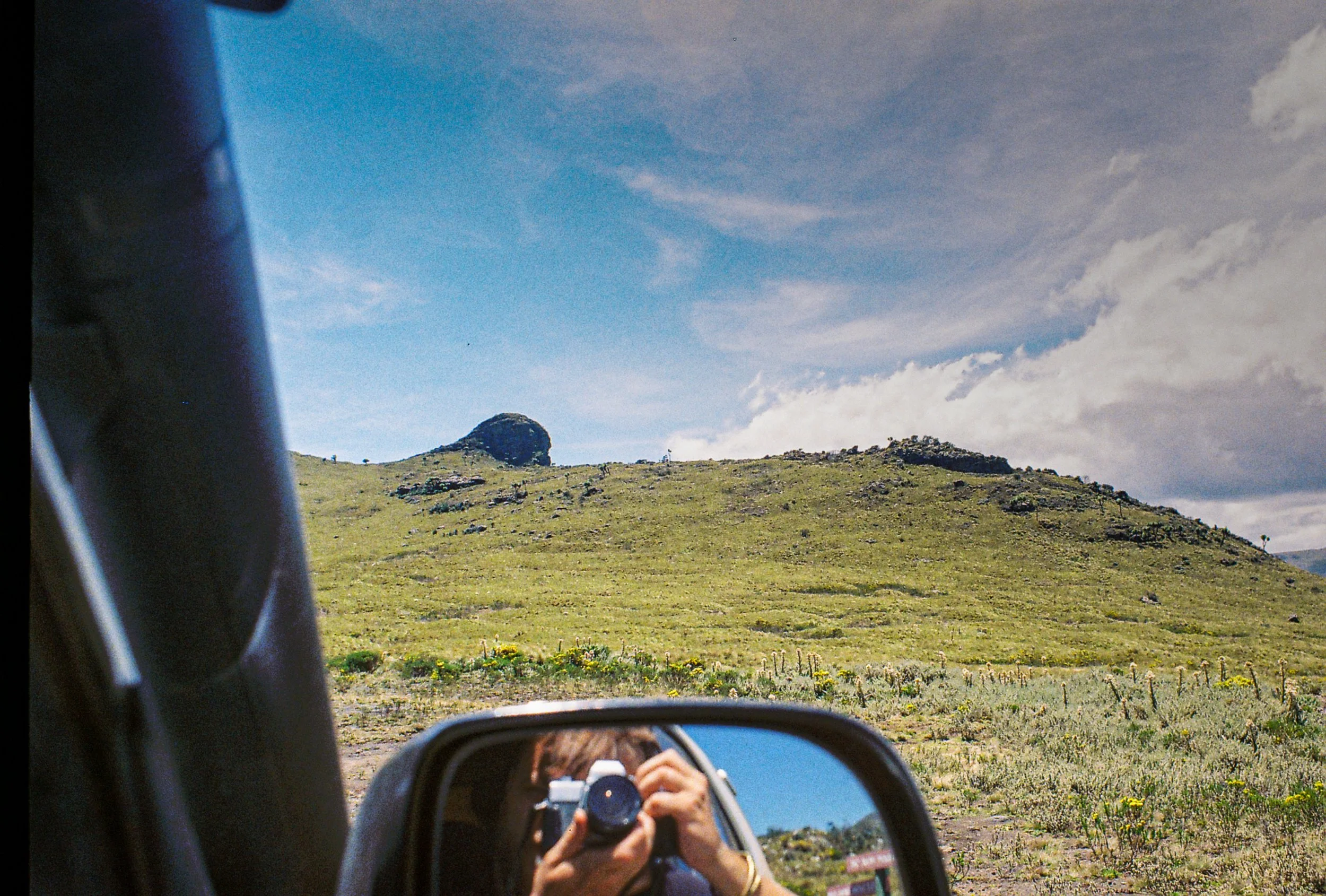 Photo of a person taking a picture of a landscape visible in the side mirror of a car. The landscape features a grassy hillside with a large rock formation, under a partly cloudy sky.
