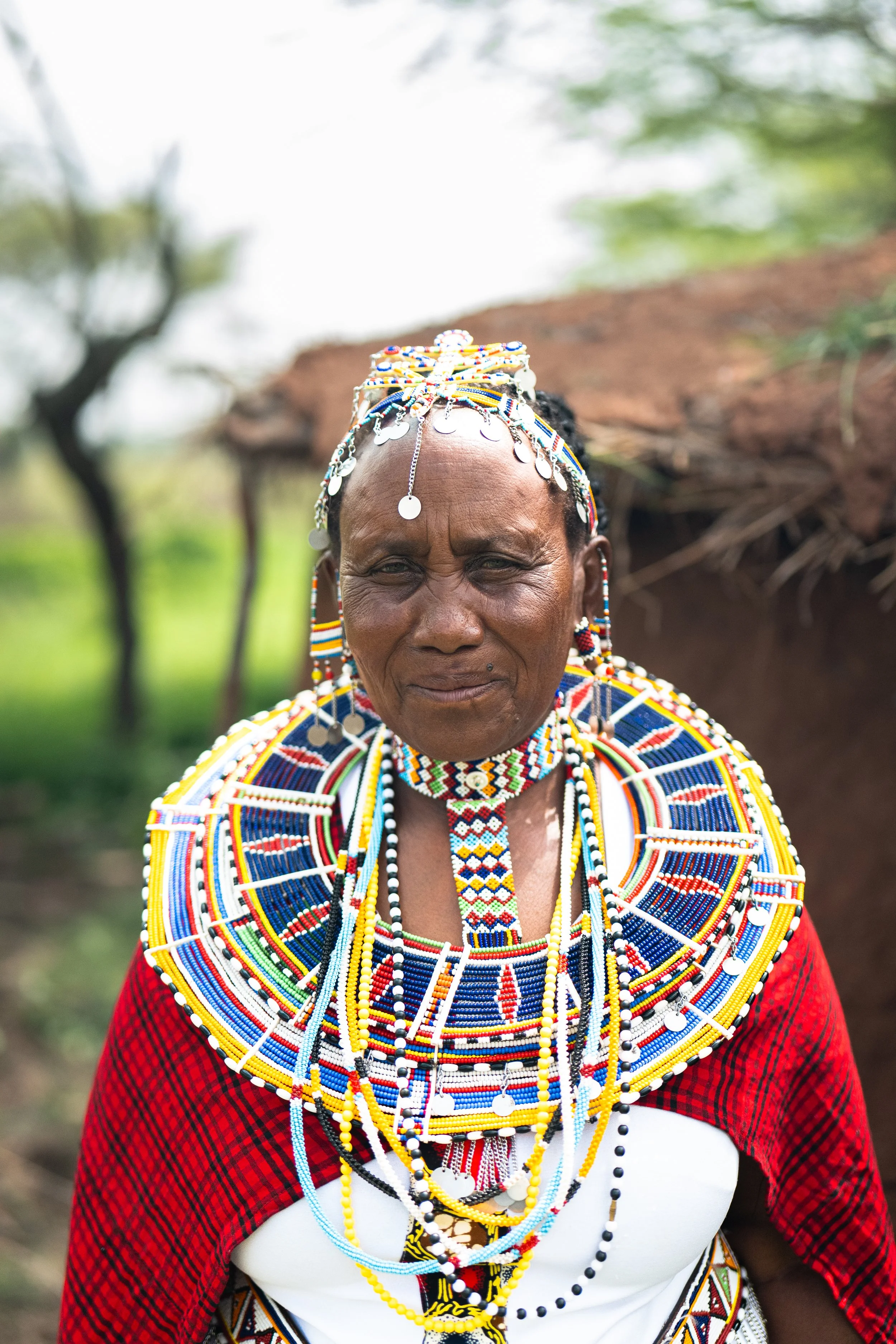 A woman wearing traditional Maasai attire, including colorful beaded jewelry, necklaces, and headpieces, standing outdoors with blurred trees and greenery in the background.