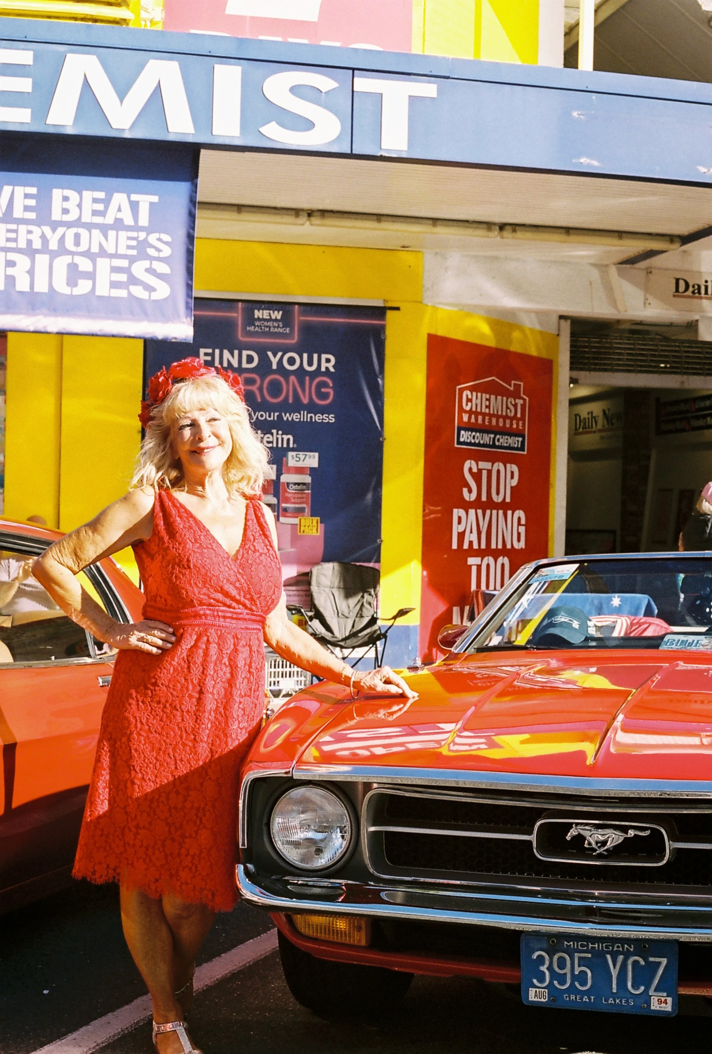 A woman in a red lace dress standing next to a red Ford Mustang with Michigan license plates at a car show.
