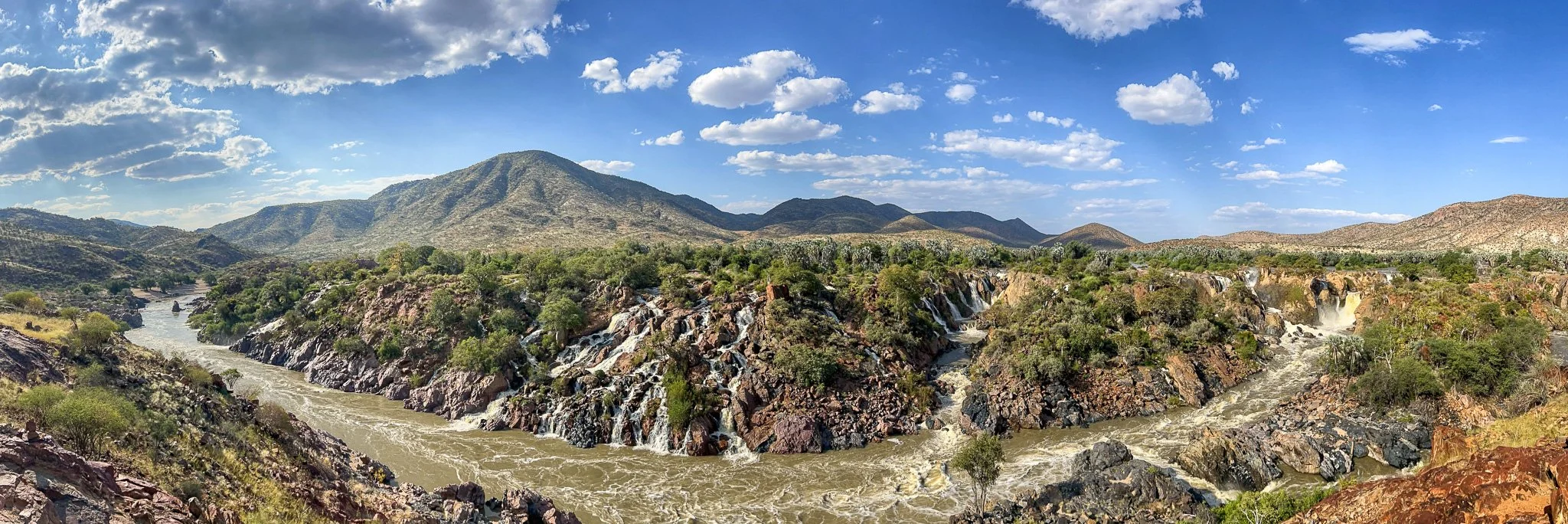 A panoramic view of a river flowing through a rocky canyon with green trees and shrubbery, surrounded by mountains under a partly cloudy sky.