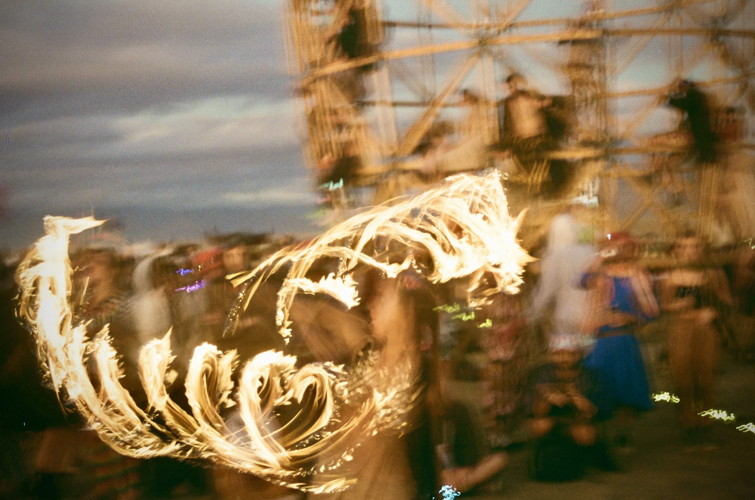 People gathered outdoors at night, around a fire with swirling orange flames, and a large metal structure in the background under dark, cloudy sky.