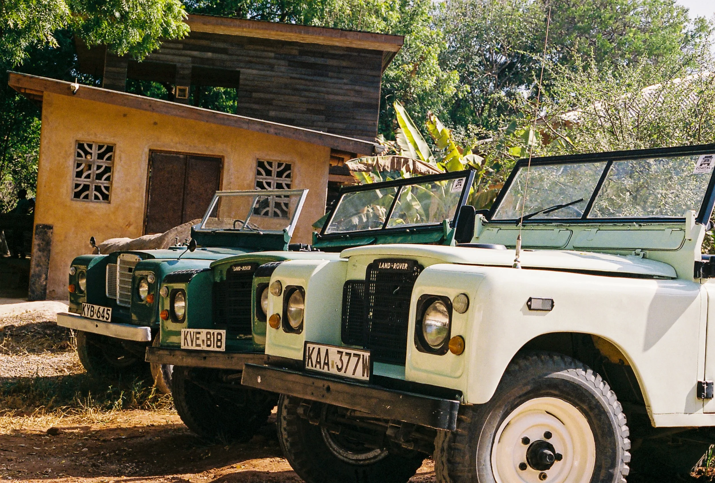 A row of vintage Land Rover vehicles parked outdoors in front of a rustic building with a wooden upper level and a yellowish lower level, surrounded by lush green trees and foliage.