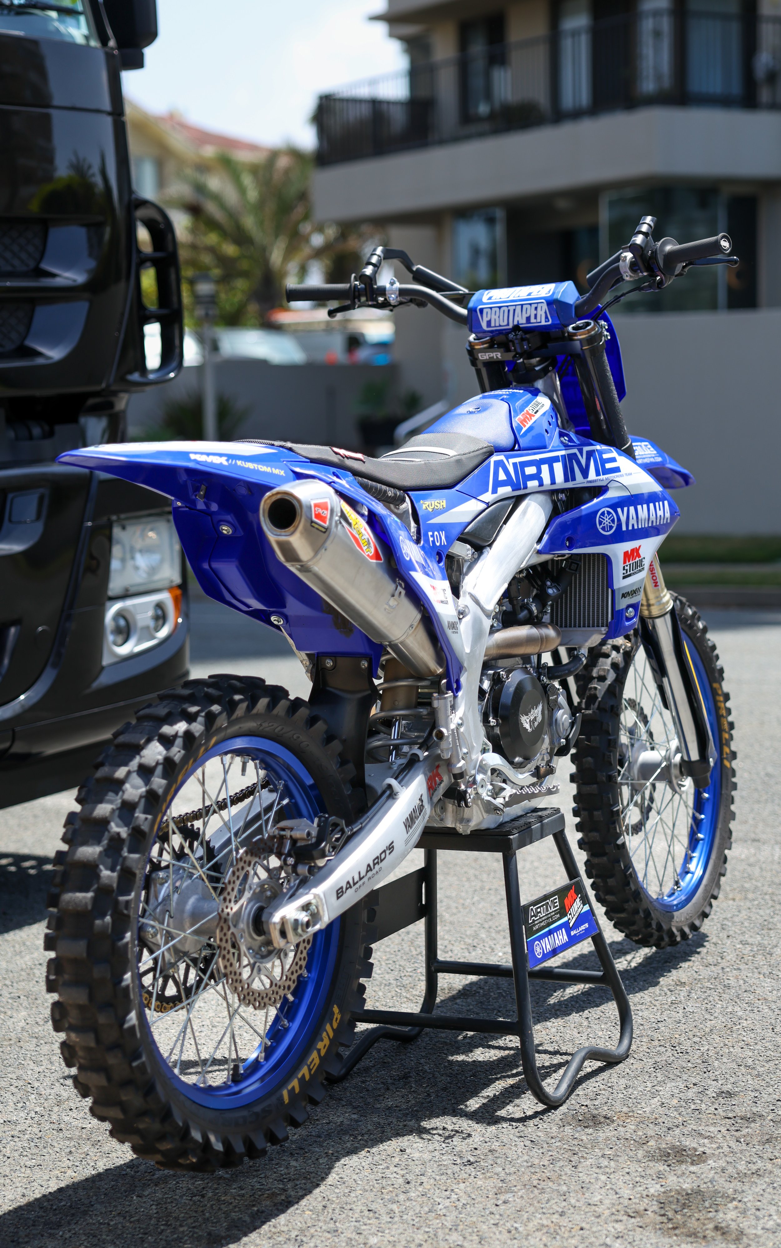 A blue Yamaha dirt bike with sponsorship logos displayed on a black stand outdoors, with a residential building and greenery in the background.