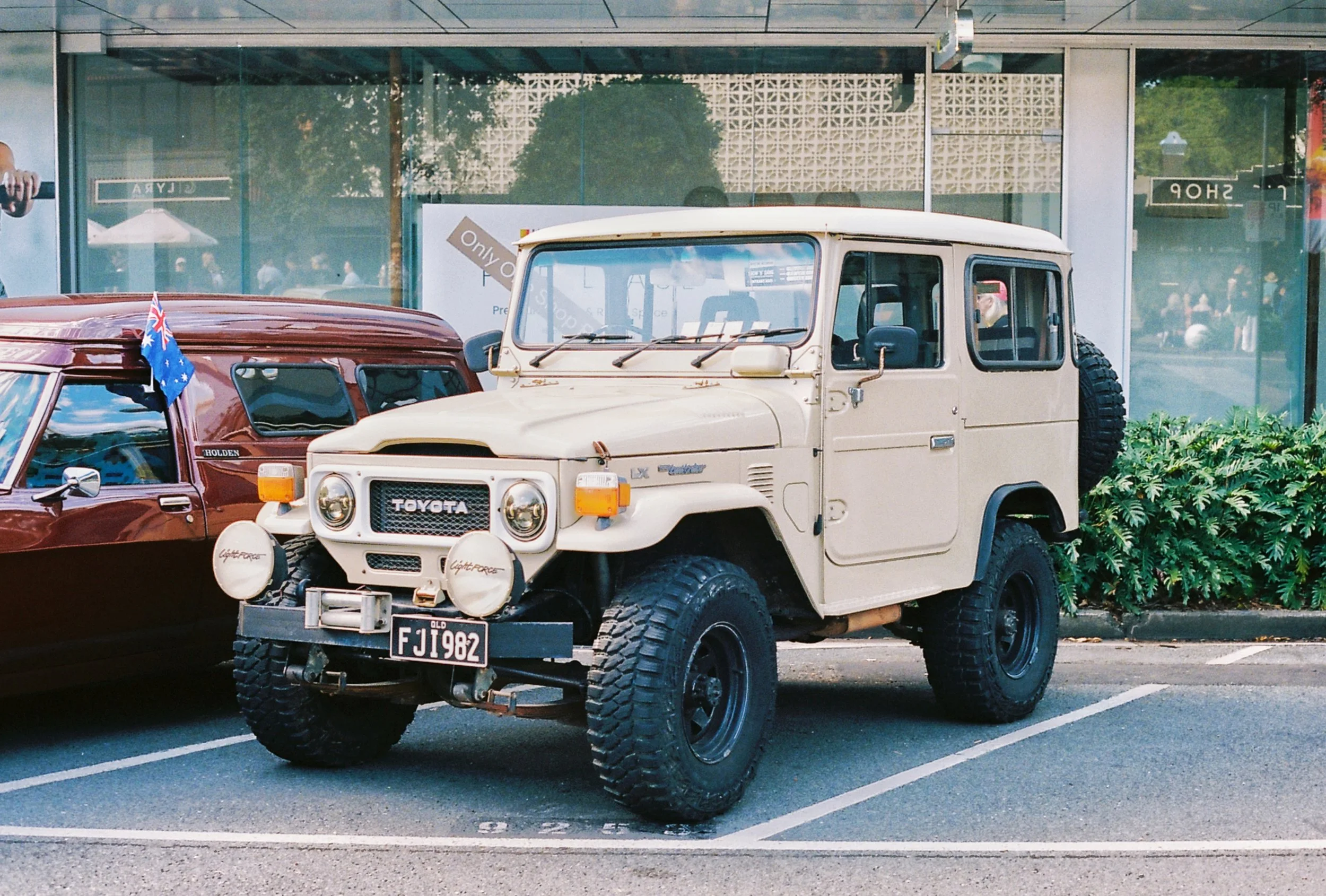 A vintage white Toyota Land Cruiser parked in a parking lot next to a maroon car with an Australian flag on its window, in front of a glass building.