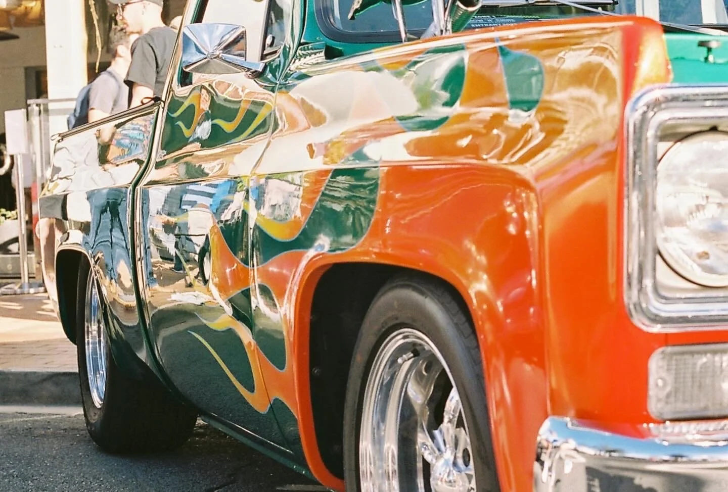 Close-up of a colorful, reflective vintage car with orange, green, and yellow paint at an outdoor event.