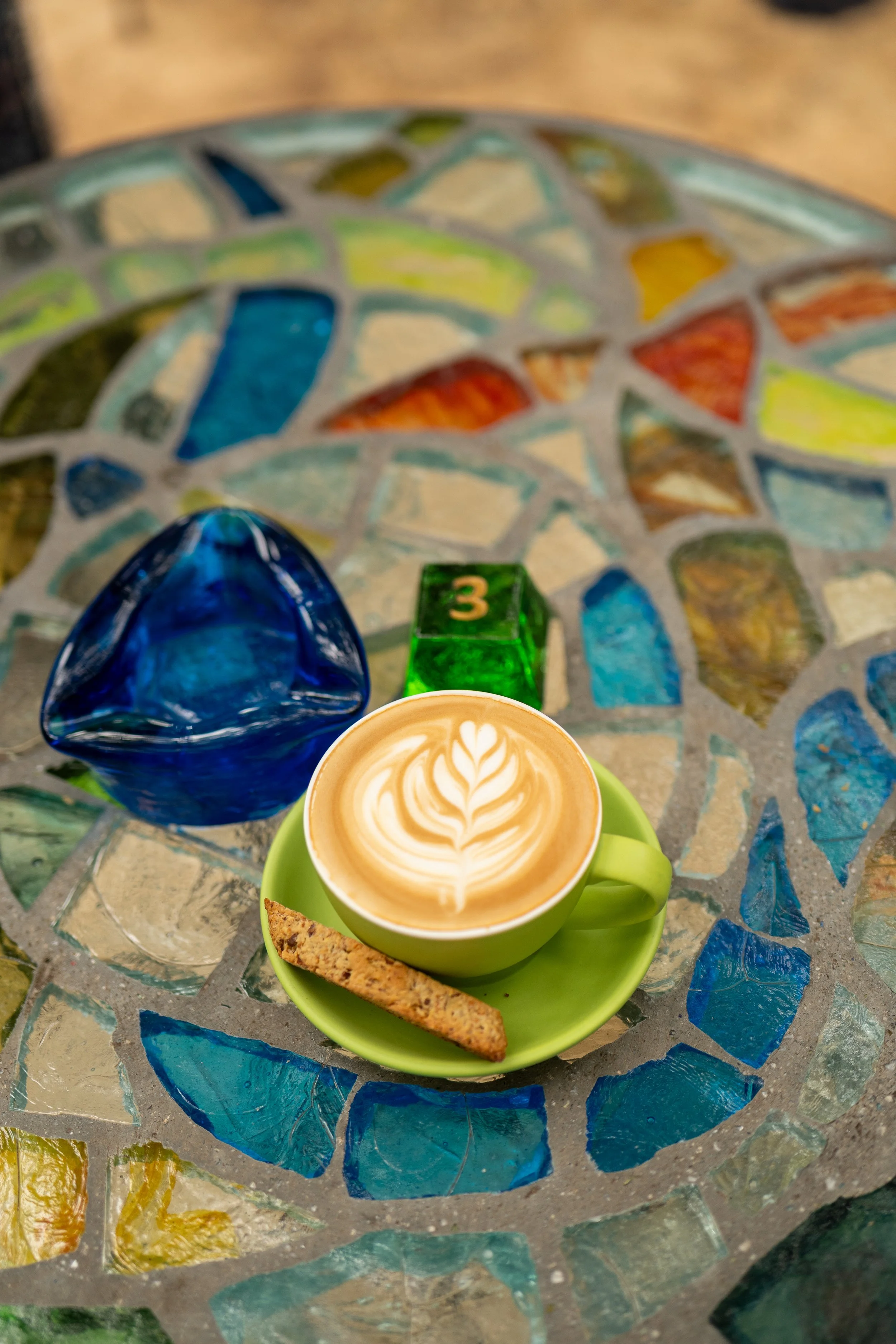 A close-up view of a colorful mosaic table with a green cup of latte art, a biscotti, a blue glass paperweight, and a small green block with the number 3, all on a glass mosaic surface.