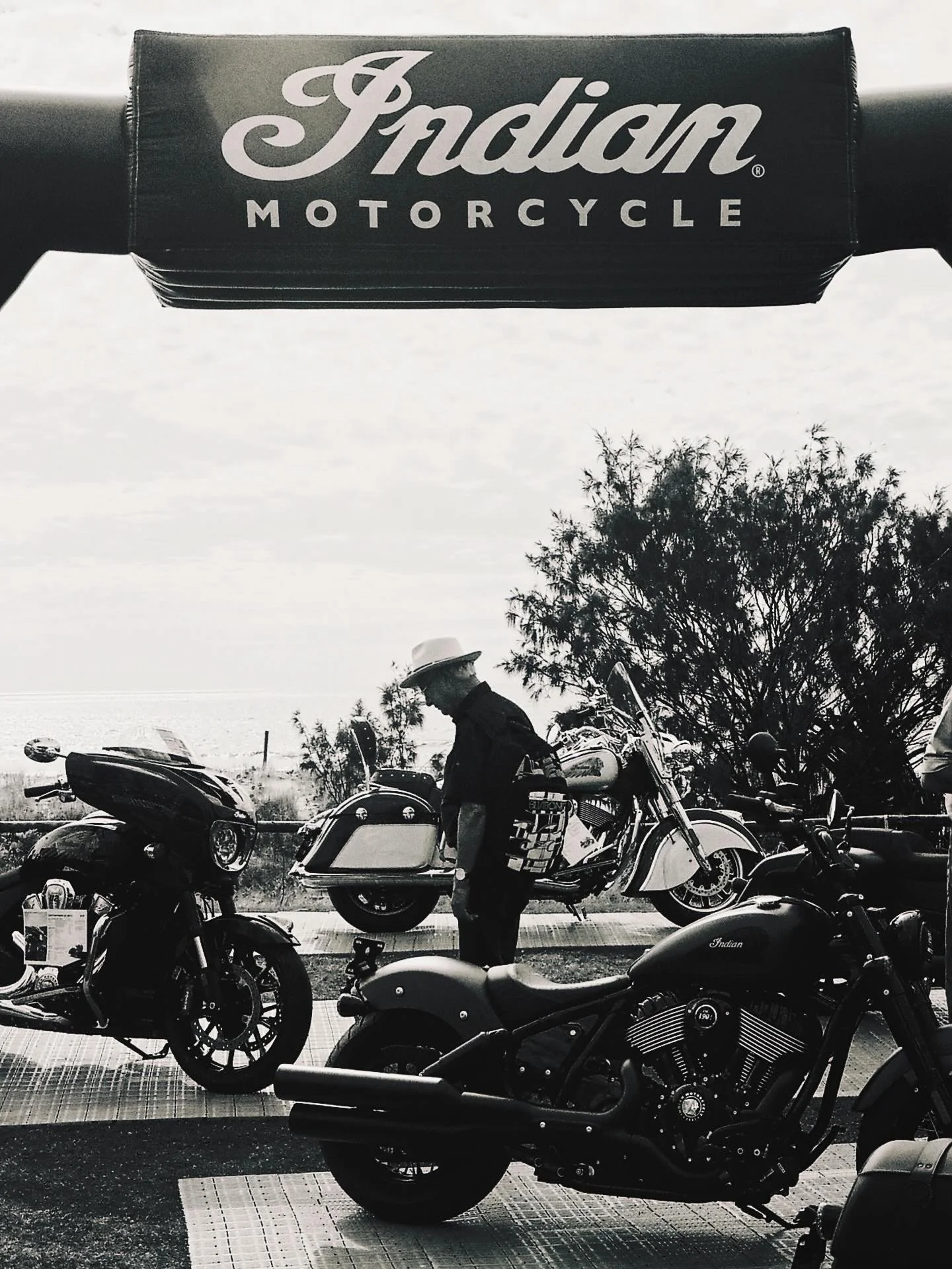 Black and white photograph of a parking lot with several motorcycles and a person standing among them. There is an Indian motorcycle in the foreground and a row of other motorcycles in the background. The person is wearing a hat and appears to be ins