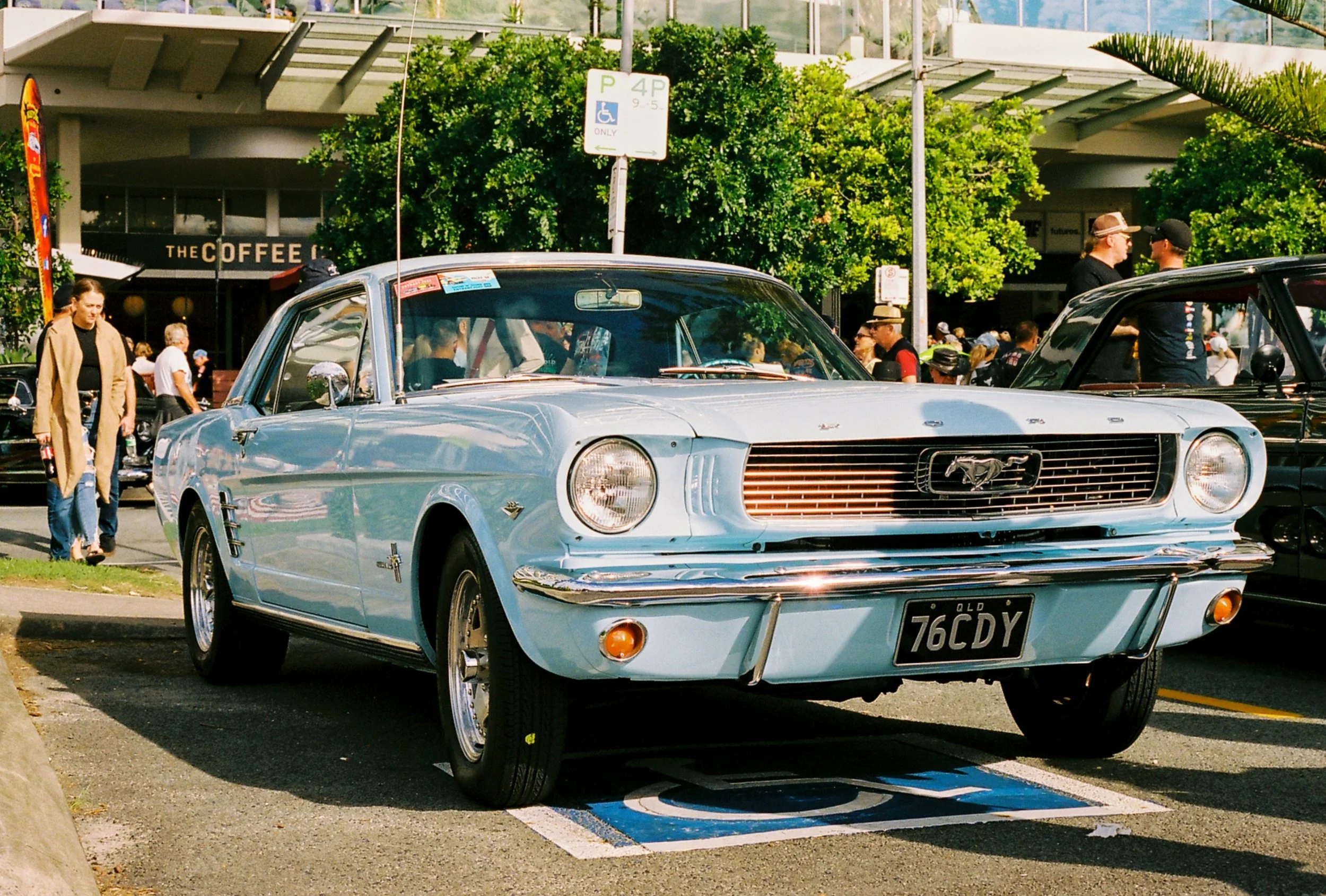 A classic light blue Ford Mustang parked in a designated parking space near a busy urban area with people walking around, trees, and buildings in the background.