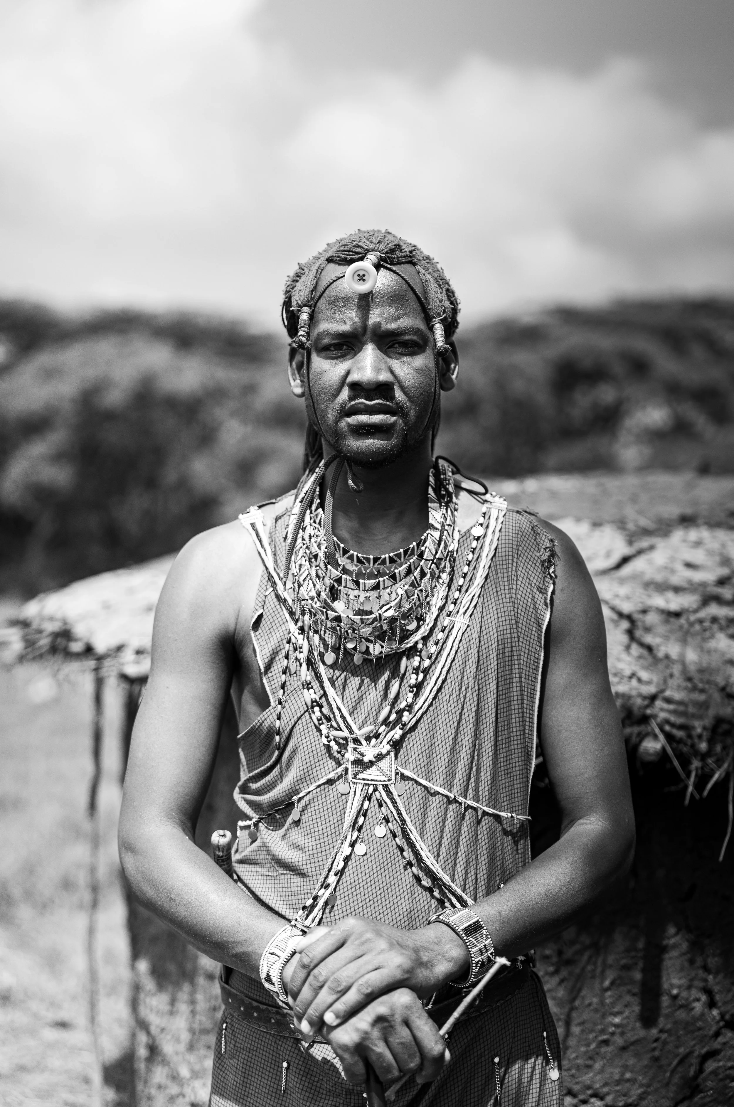 A portrait of a man from an indigenous community, dressed in traditional attire with layered necklaces and beads, standing outdoors under cloudy sky.