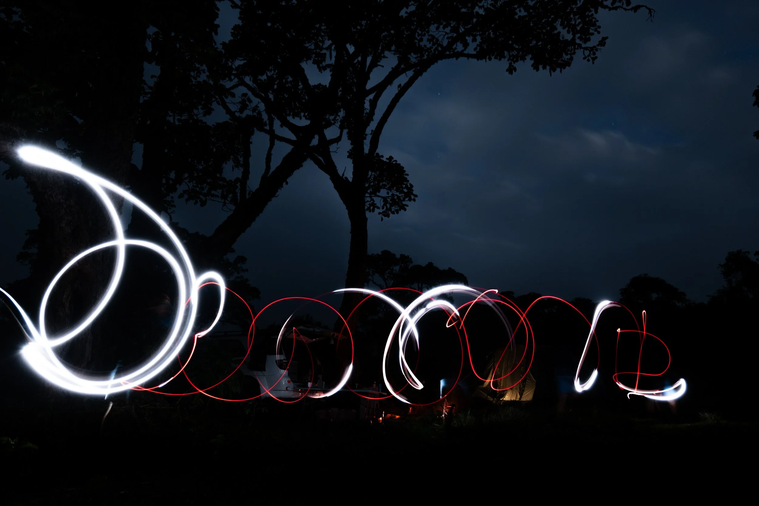 Nighttime outdoor scene with light trail patterns in white and red, silhouette of trees, dark sky with some clouds, and a faint star.