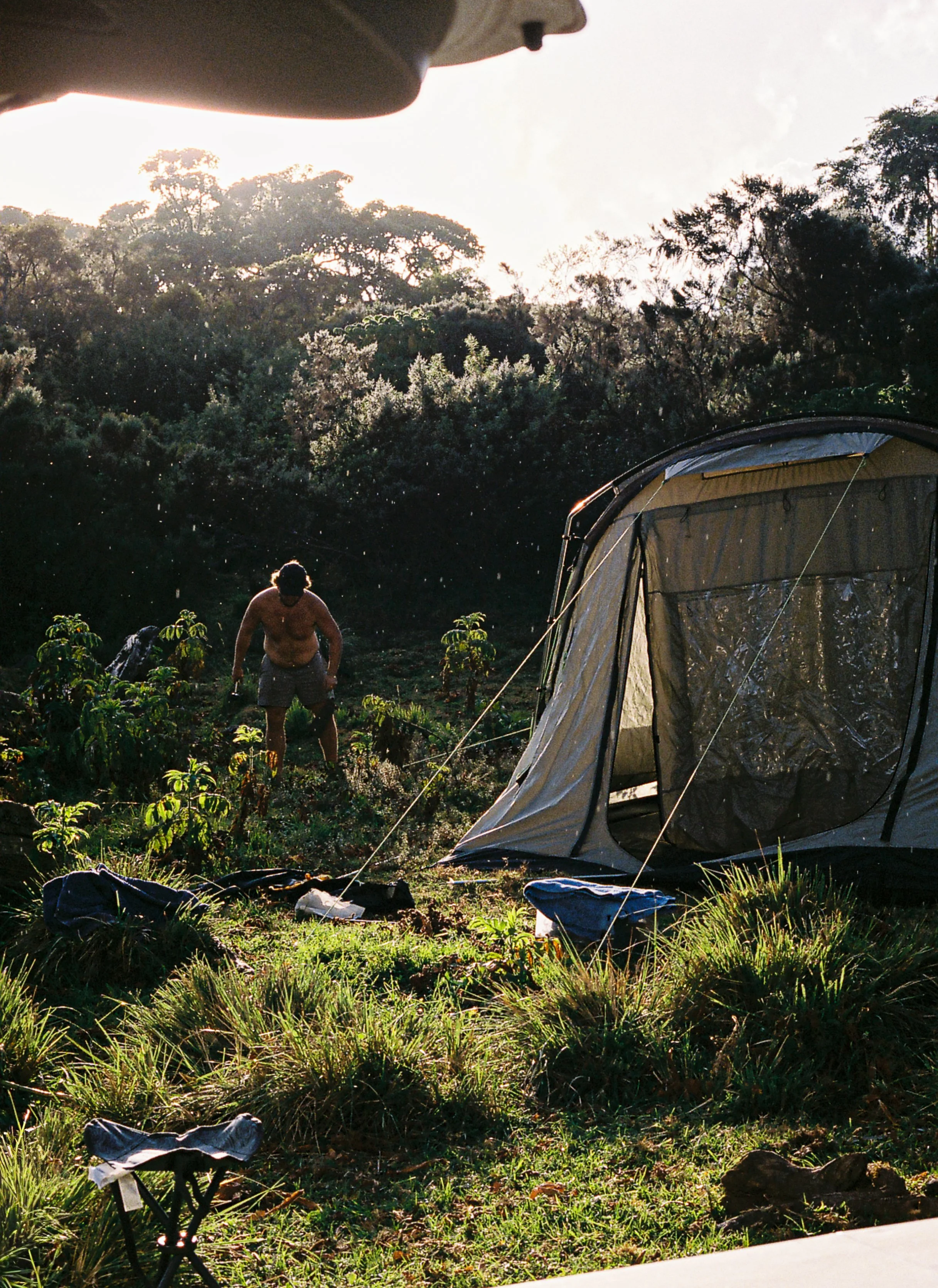A person without a shirt and wearing shorts is working in a garden with small plants, near a camping tent set up outdoors. The scene is backlit by the sun, with lush green trees in the background.