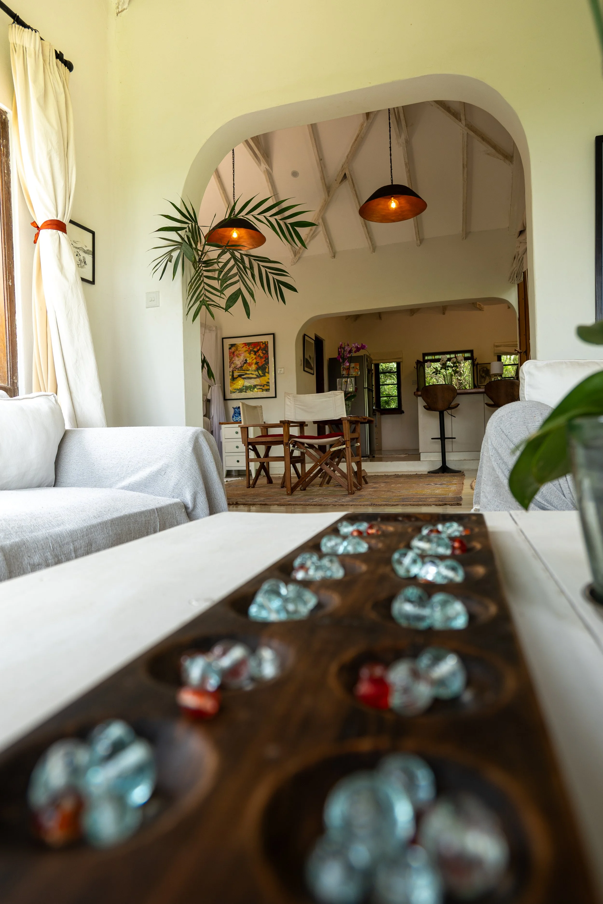 Living room with white couches, a wooden dining table with chairs, artwork on the walls, and a small kitchen area with bar stools and windows in the background.