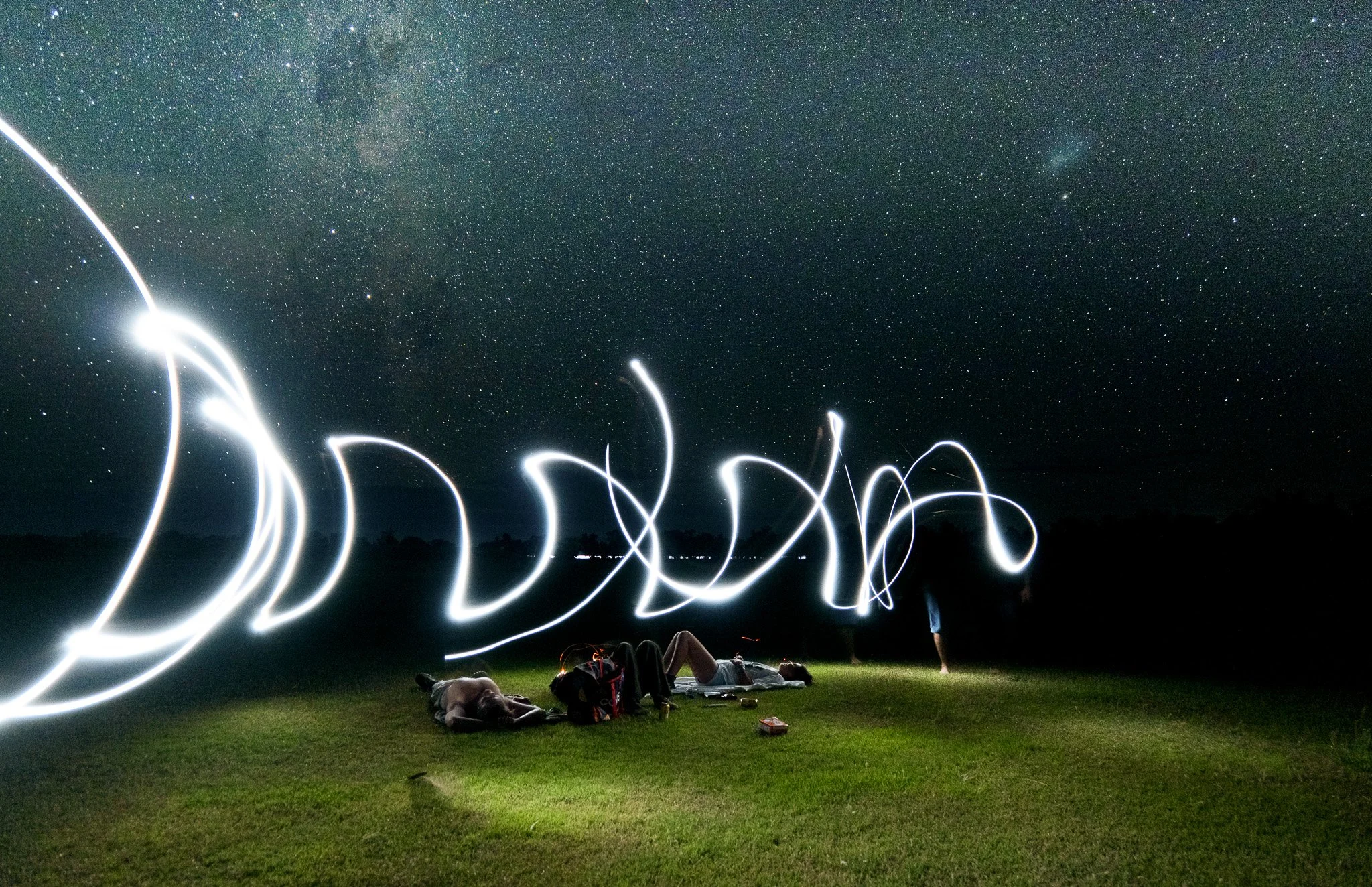 People sitting and lying on a grassy field at night, while long-exposure photography creates light trails from flashlights or sparklers, with a starry sky overhead.
