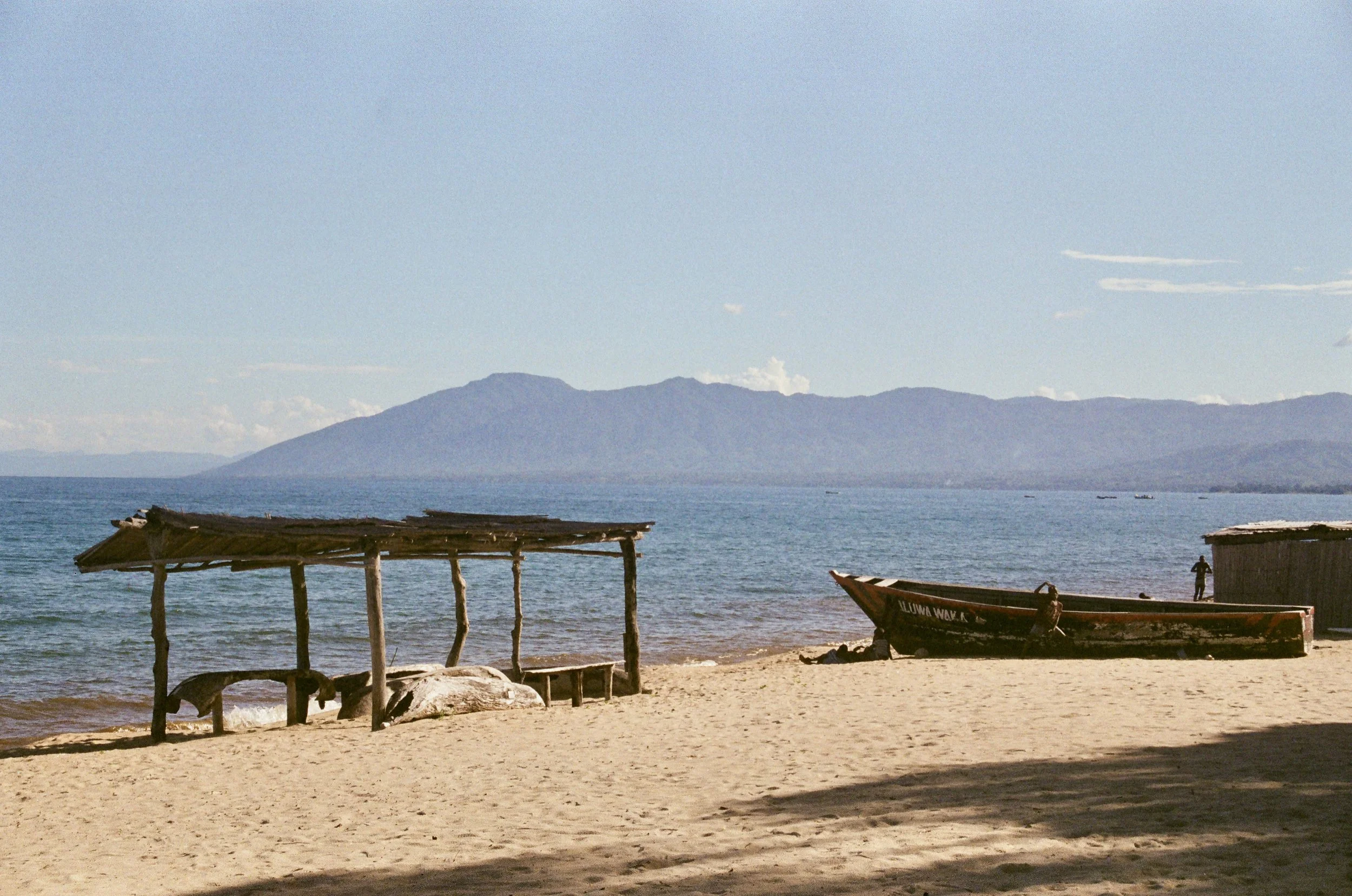 Beach scene with wooden structures and boats near the shore, mountains in the background under a clear sky.