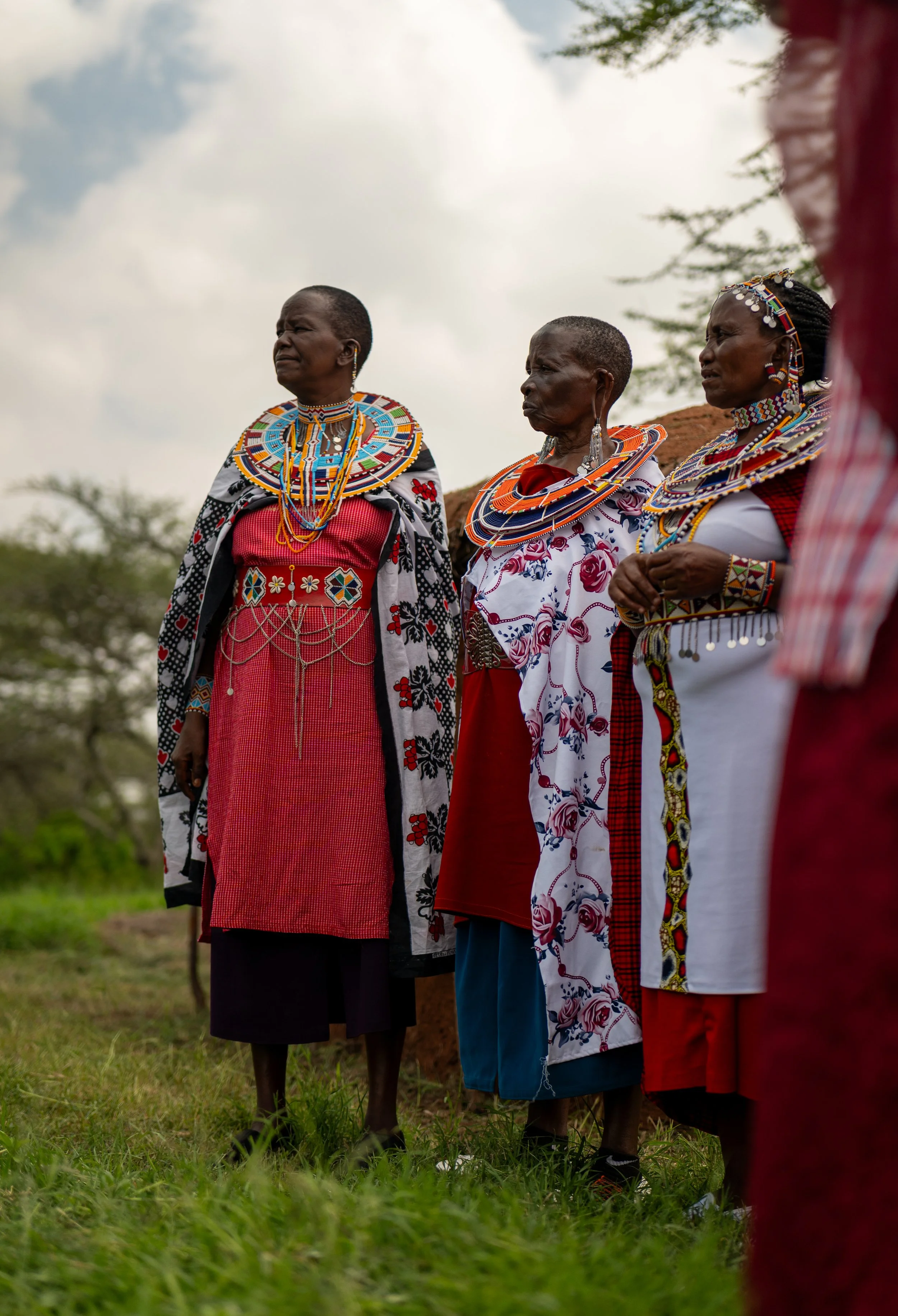 Three women dressed in traditional, colorful Maasai attire and jewelry standing outdoors with cloudy sky and trees in the background.