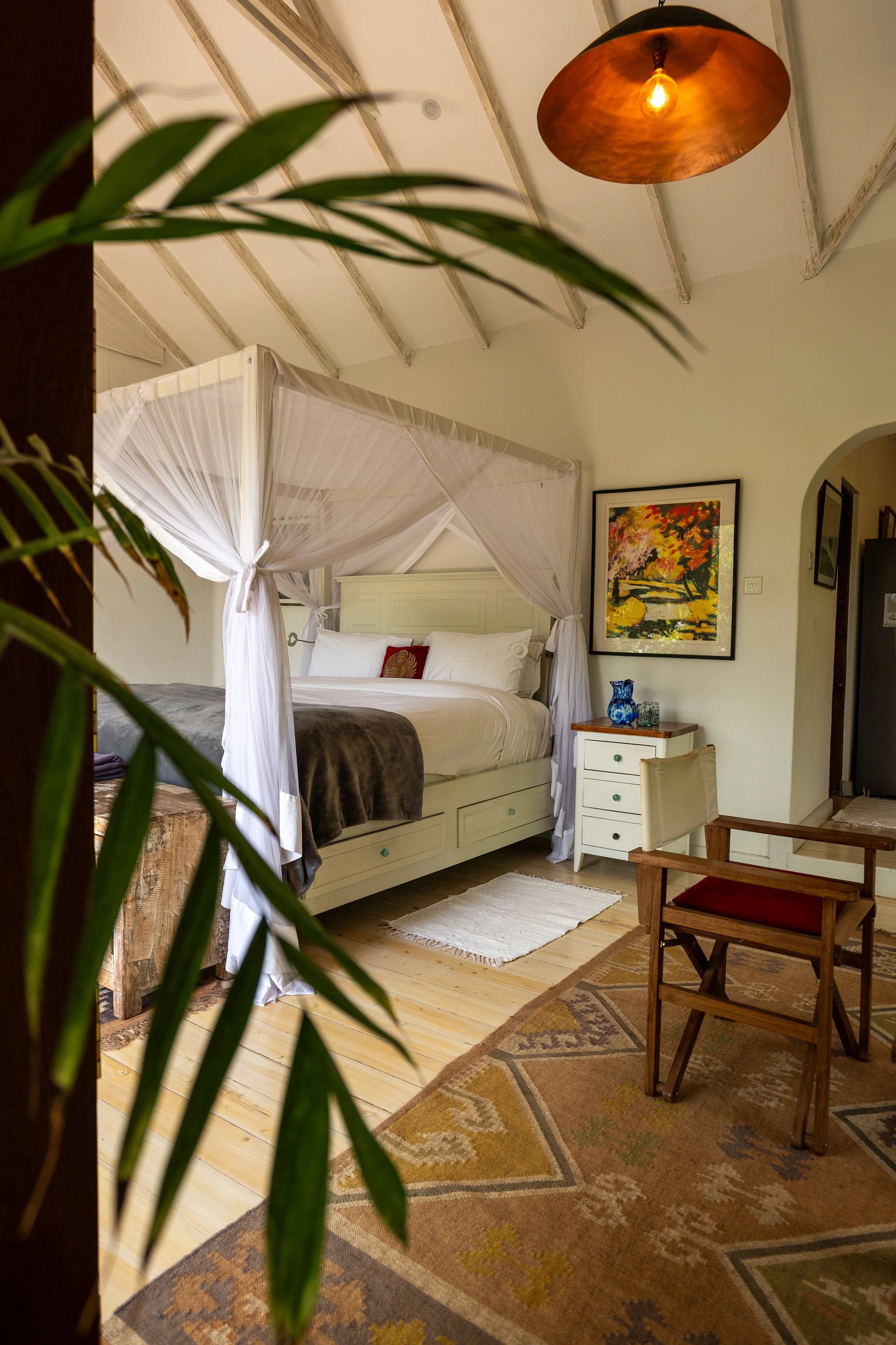 A cozy bedroom with a white four-poster bed, white canopy curtains, and white bedding. There is a colorful framed painting on the wall, a small wooden side table with a blue vase, and a wooden table with a red-cushioned chair. The area rug has an ear