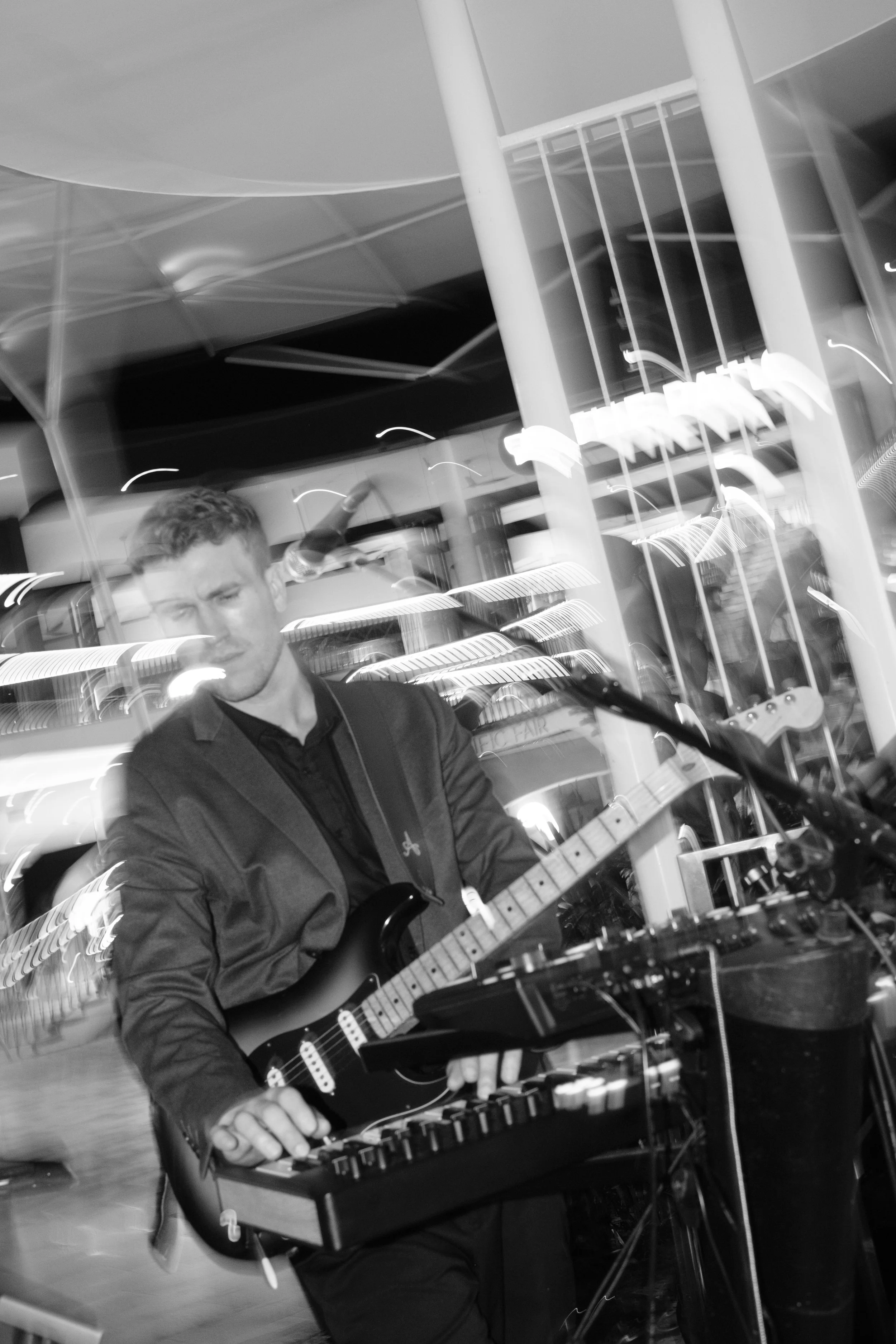 A young man playing an electric guitar connected to music equipment in an indoor space with modern lighting and reflective surfaces.