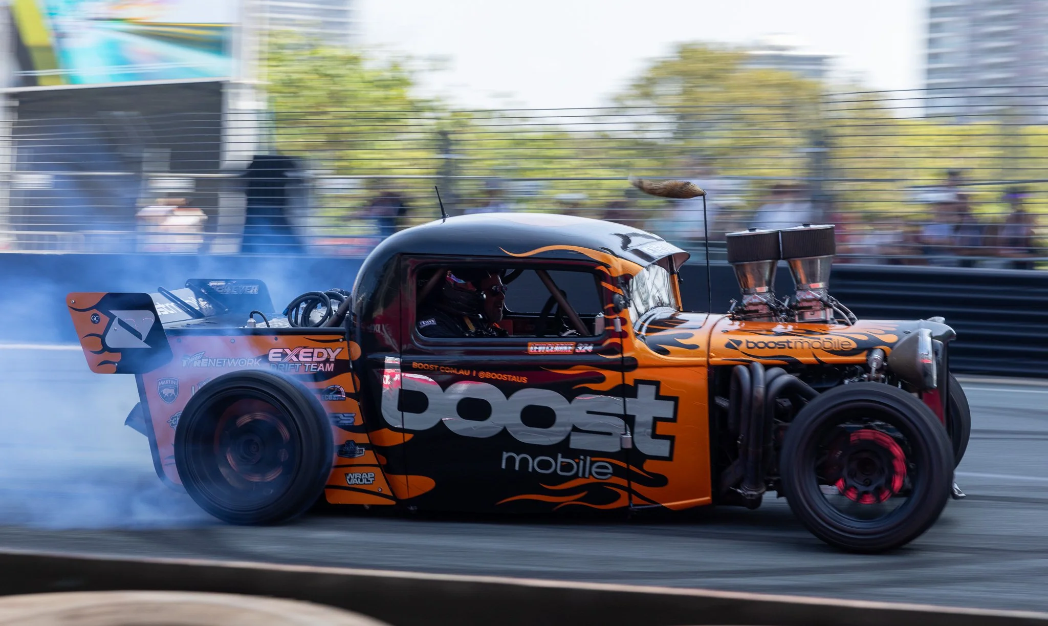 A drag racing car with an orange and black flame design speeds down the track, creating smoke behind the tires, with a blurred crowd and trees in the background.