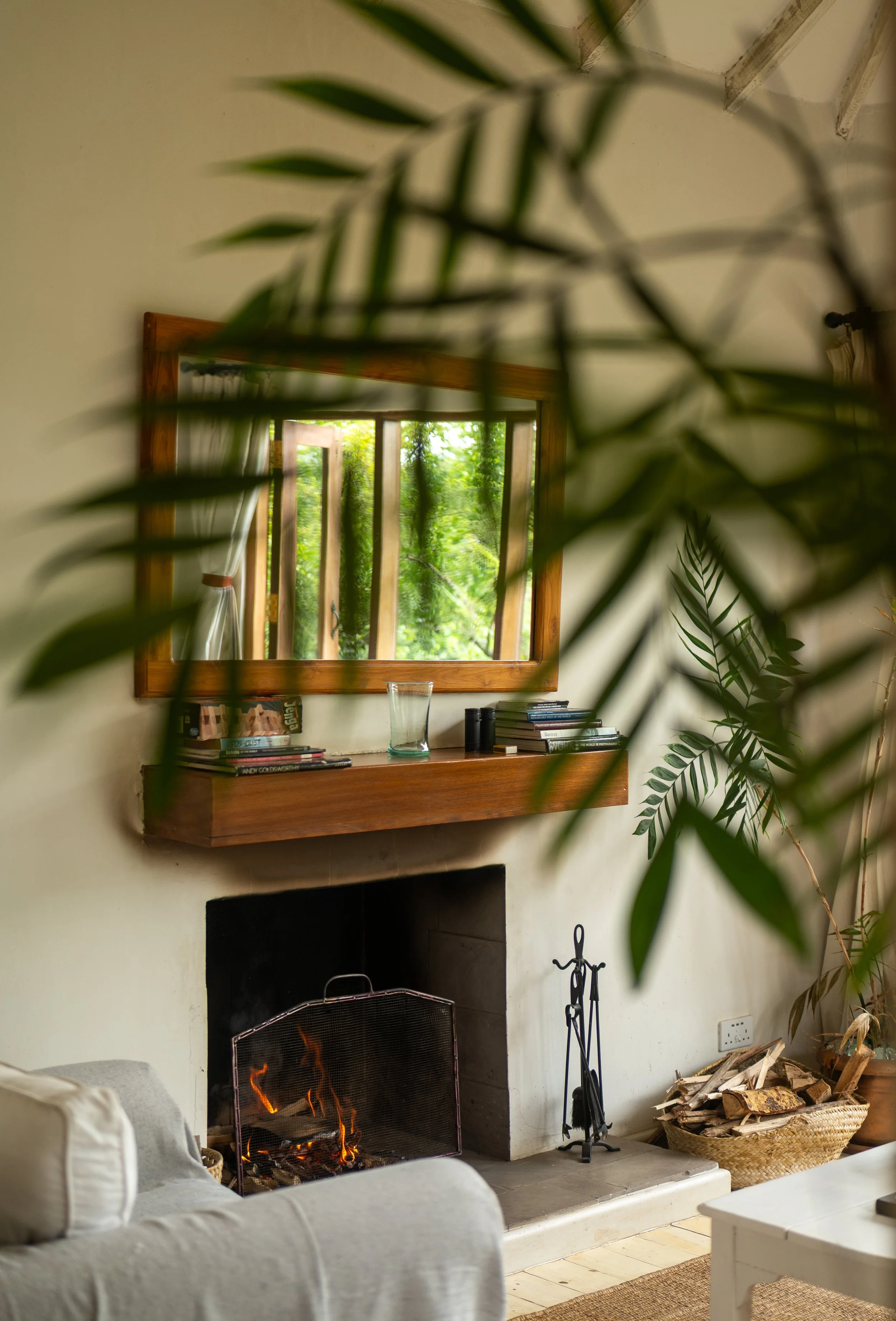 Living room with a lit fireplace, a wall-mounted mirror, a wooden mantel with books and a glass, and a basket of firewood. Green foliage is visible outside through an open window.