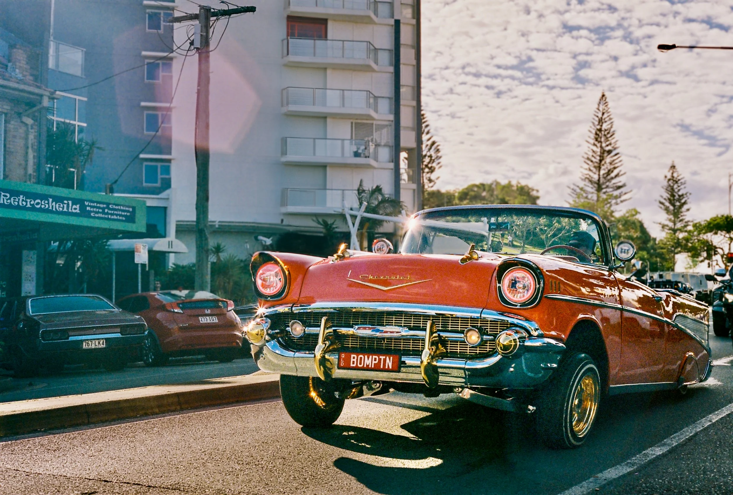 A vintage red Chevrolet convertible parked on a street, with a sign reading 'Retrosheila' in the background, residential buildings, trees, and a cloudy sky.