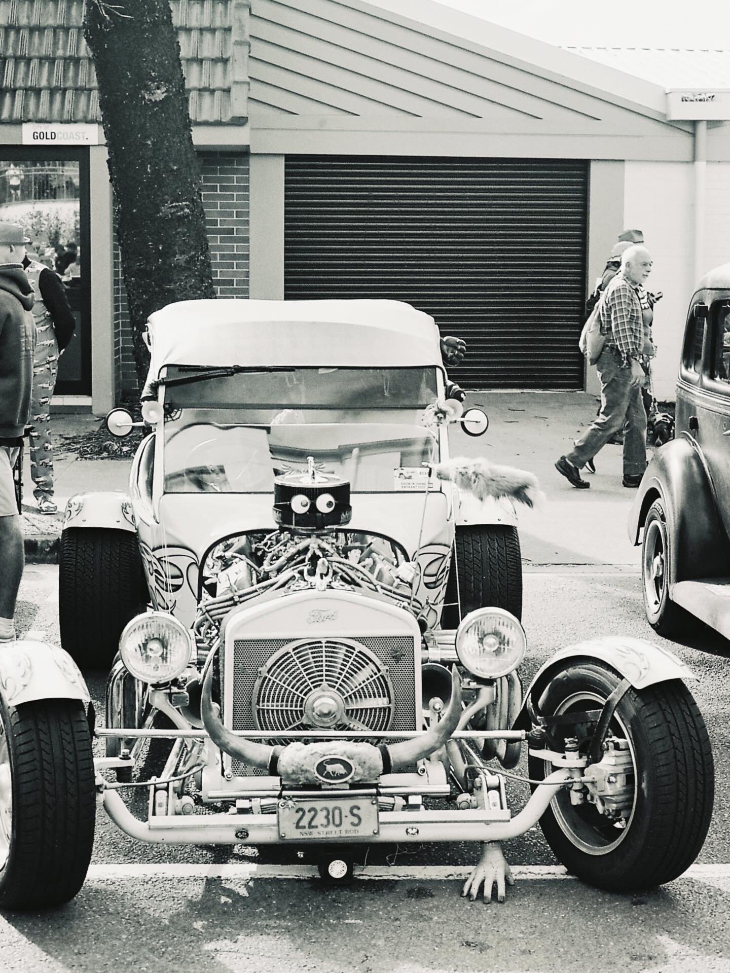 A vintage car with its hood open reveals the engine, parked on a street with people walking by. One person is walking with a backpack, and other vintage cars can be seen around.