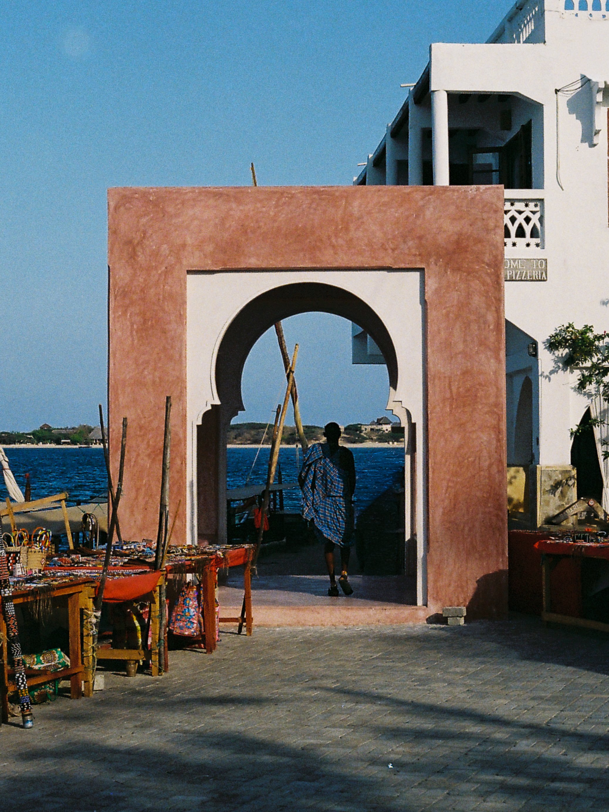 A person walking through a pink and white archway near a body of water, with vendors selling jewelry on the left side and a partly cloudy sky in the background.