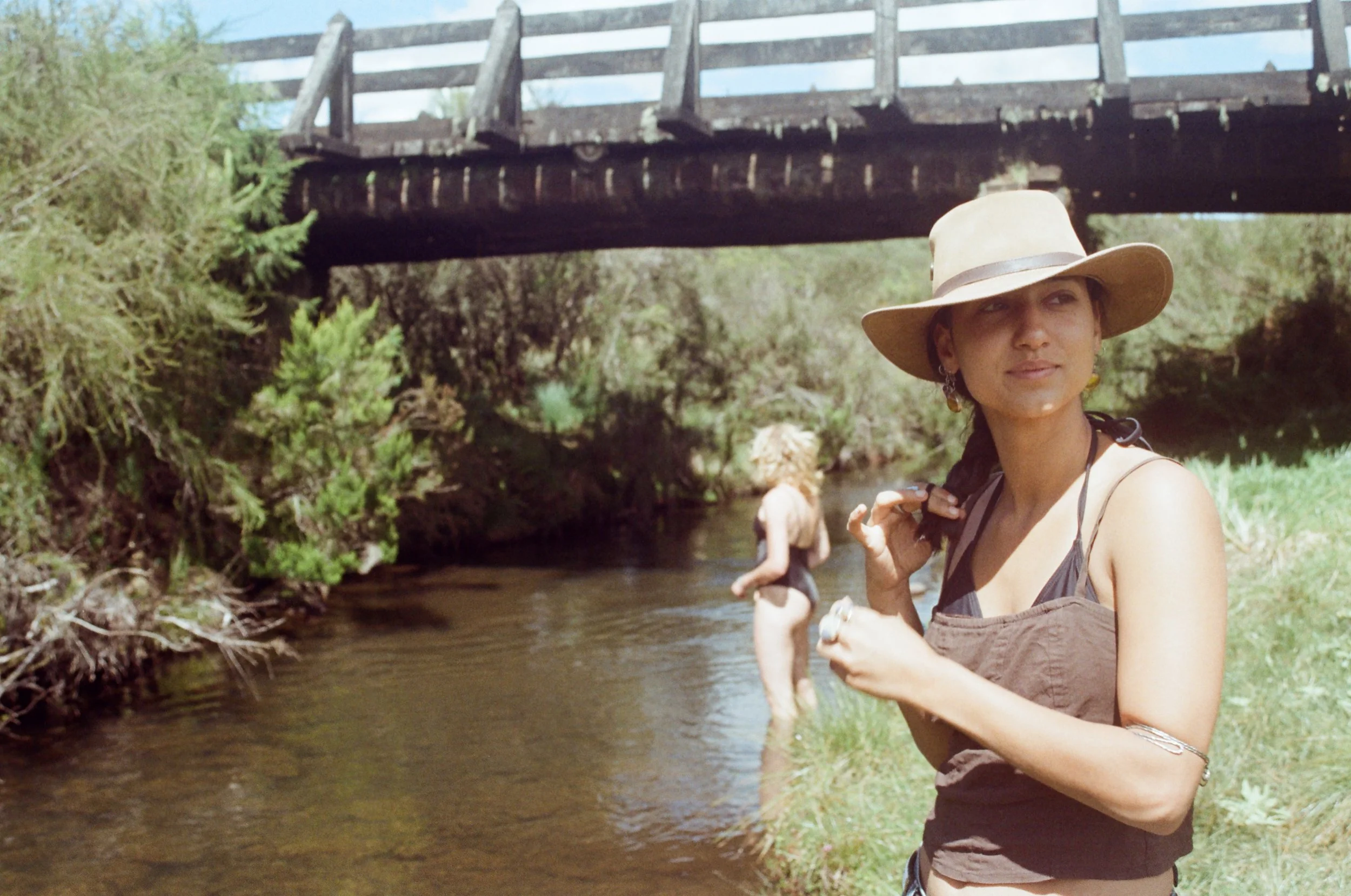A woman in a wide-brimmed hat standing by a stream, with other people in the water and a wooden bridge overhead, surrounded by lush green trees and bushes.