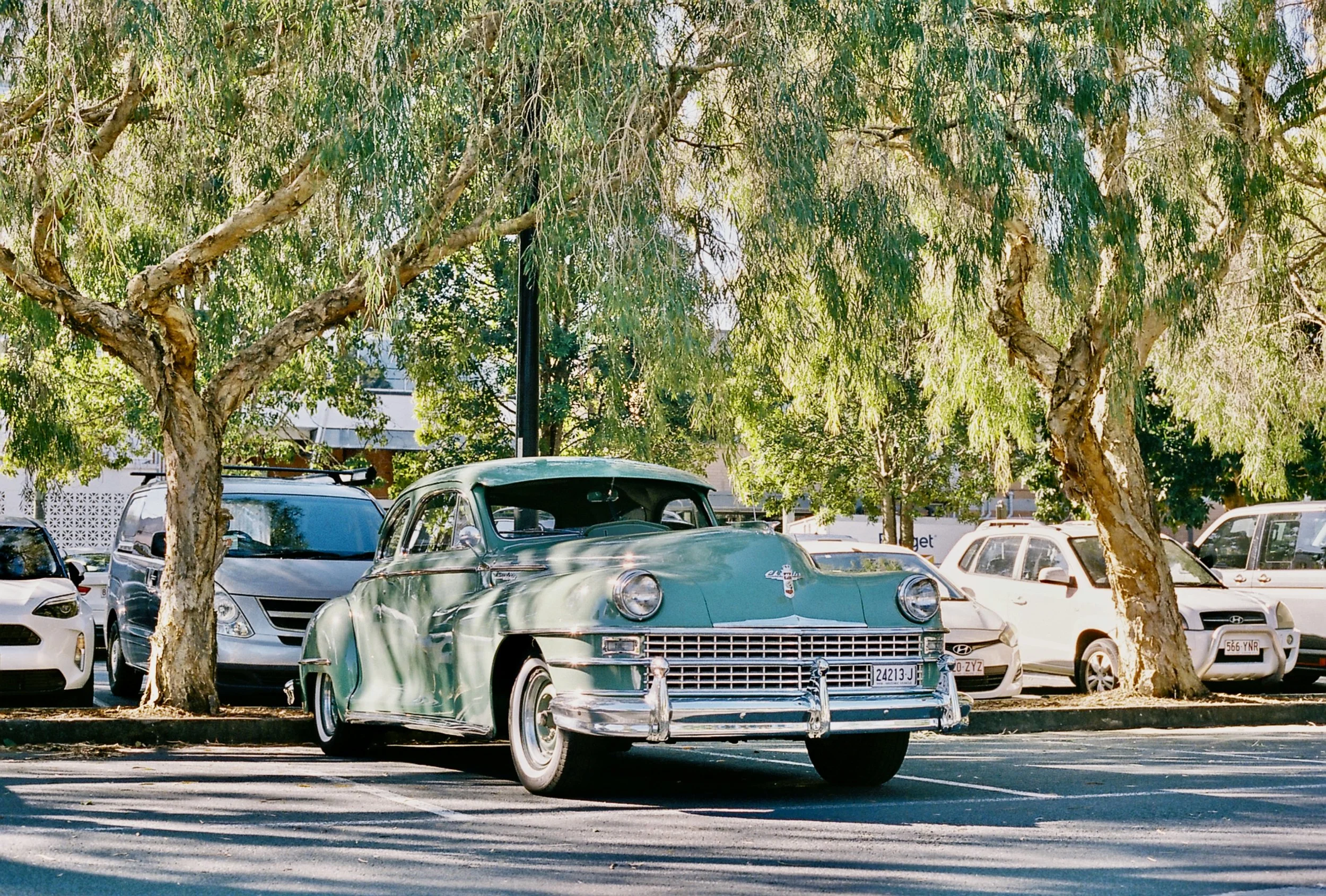 A vintage teal-colored car parked under tall green trees in a parking lot, with other modern cars nearby.