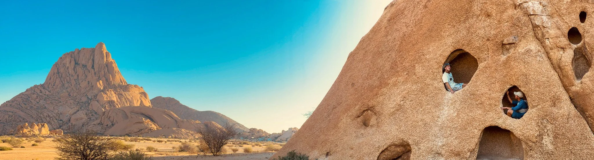People sitting inside and near large carved rock formations in a desert landscape with mountains in the background under a clear blue sky.