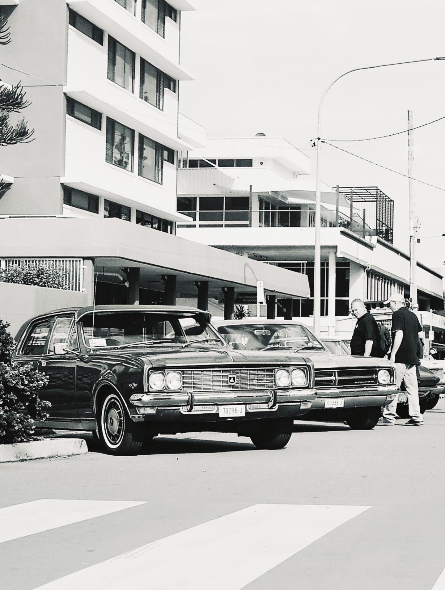 Black and white photo of a vintage car parked in front of a modern multi-story building with large windows, and two men walking past the car.