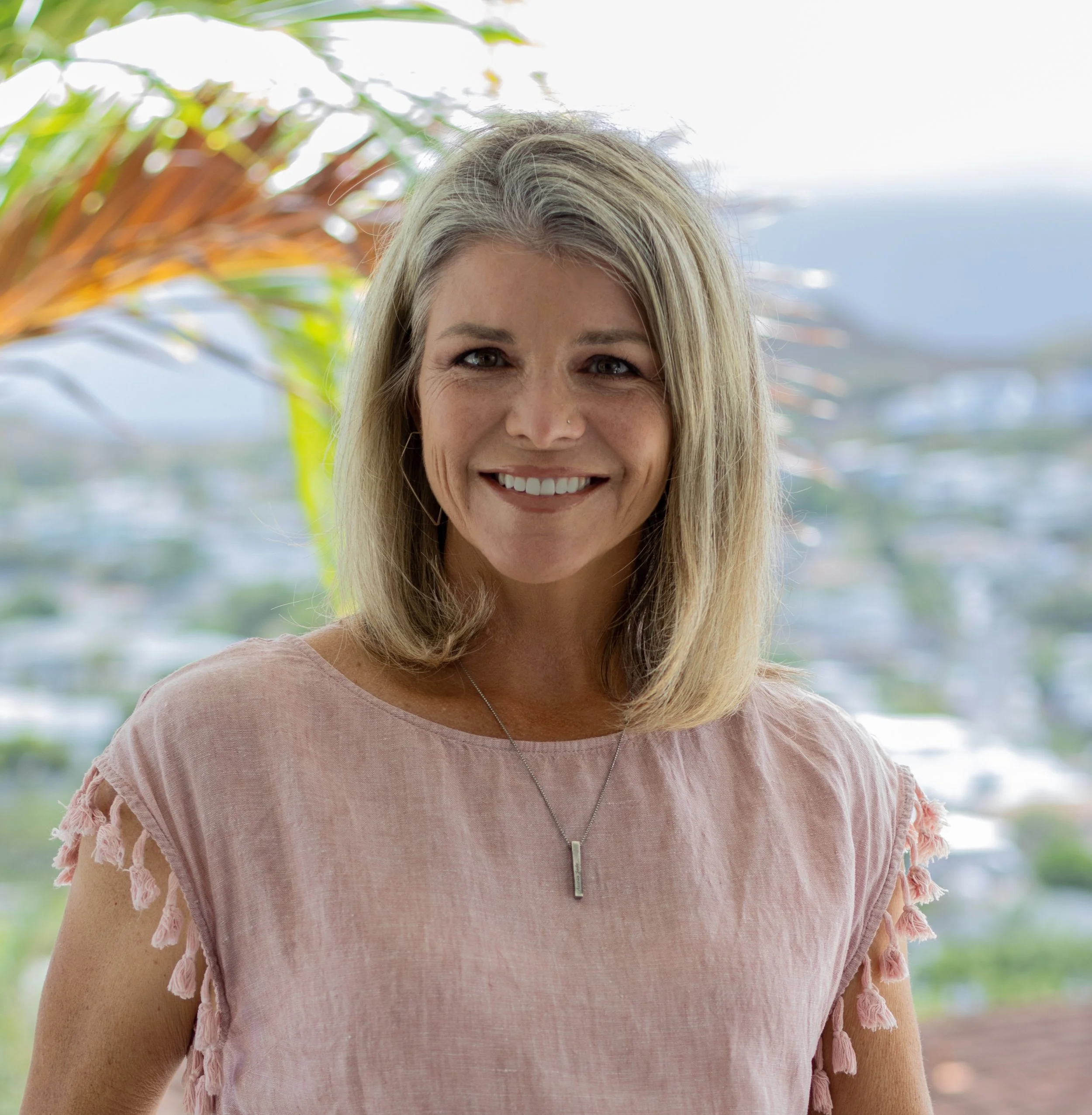 A smiling woman with blonde hair wearing a pink top with tassel details on the sleeves, standing outdoors with a blurred cityscape and palm leaves in the background.