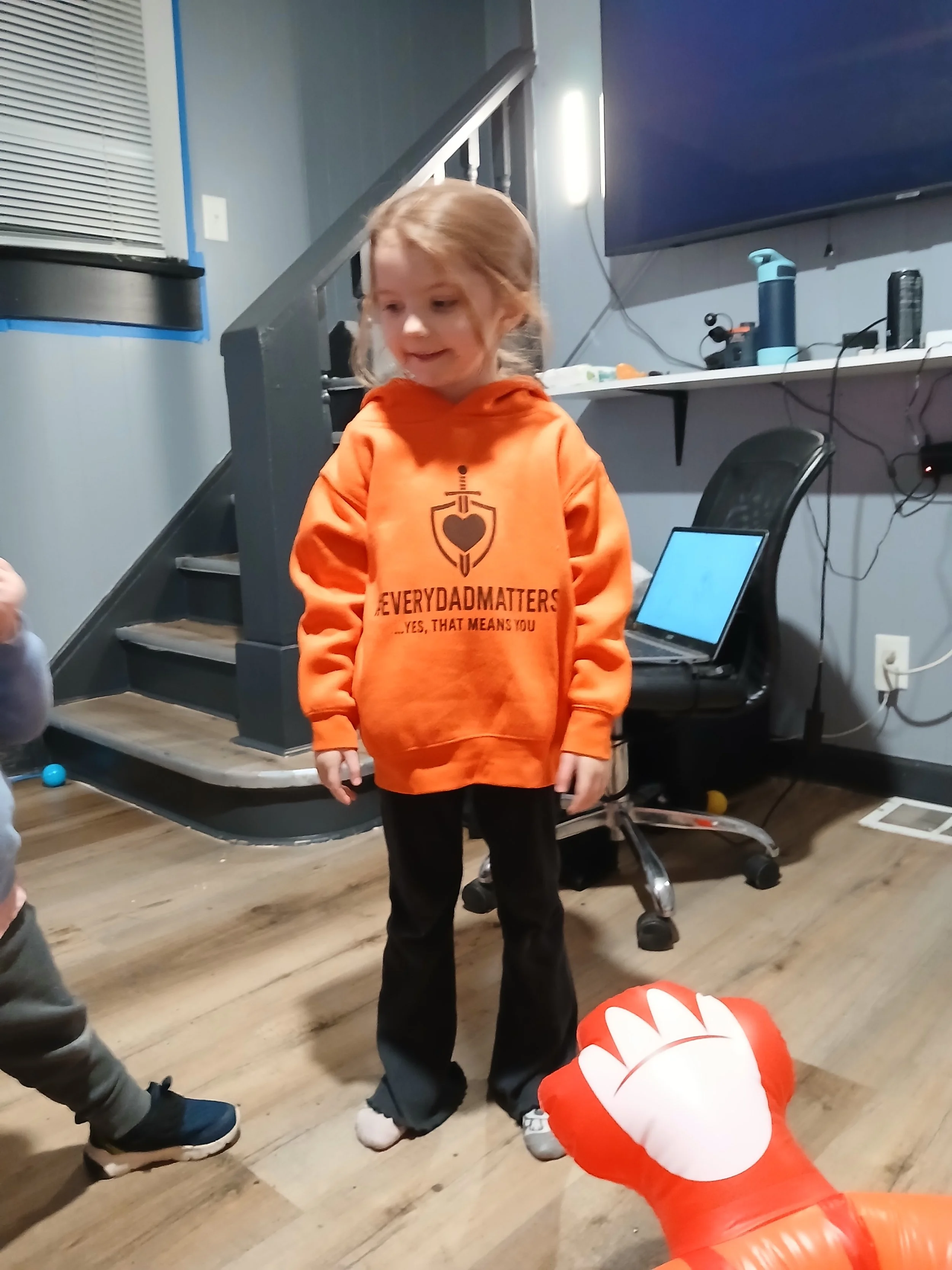 A young girl with red hair standing in a room, wearing an orange hoodie with black pants. There is a large red and white inflatable hand in the foreground and part of another child's arm on the left.