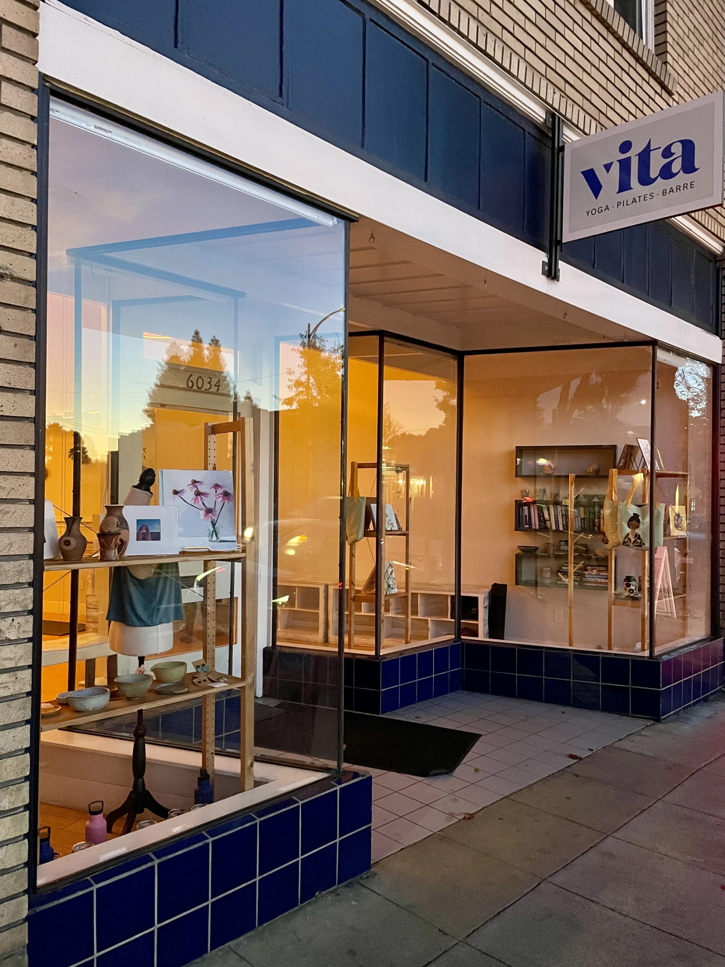 The storefront of a small shop named 'Vita' with a sign indicating it offers yoga, Pilates, and barre classes. The large windows reveal inside displays of pottery, books, and artwork with warm lighting, and the exterior features dark blue tiles and a sidewalk outside.