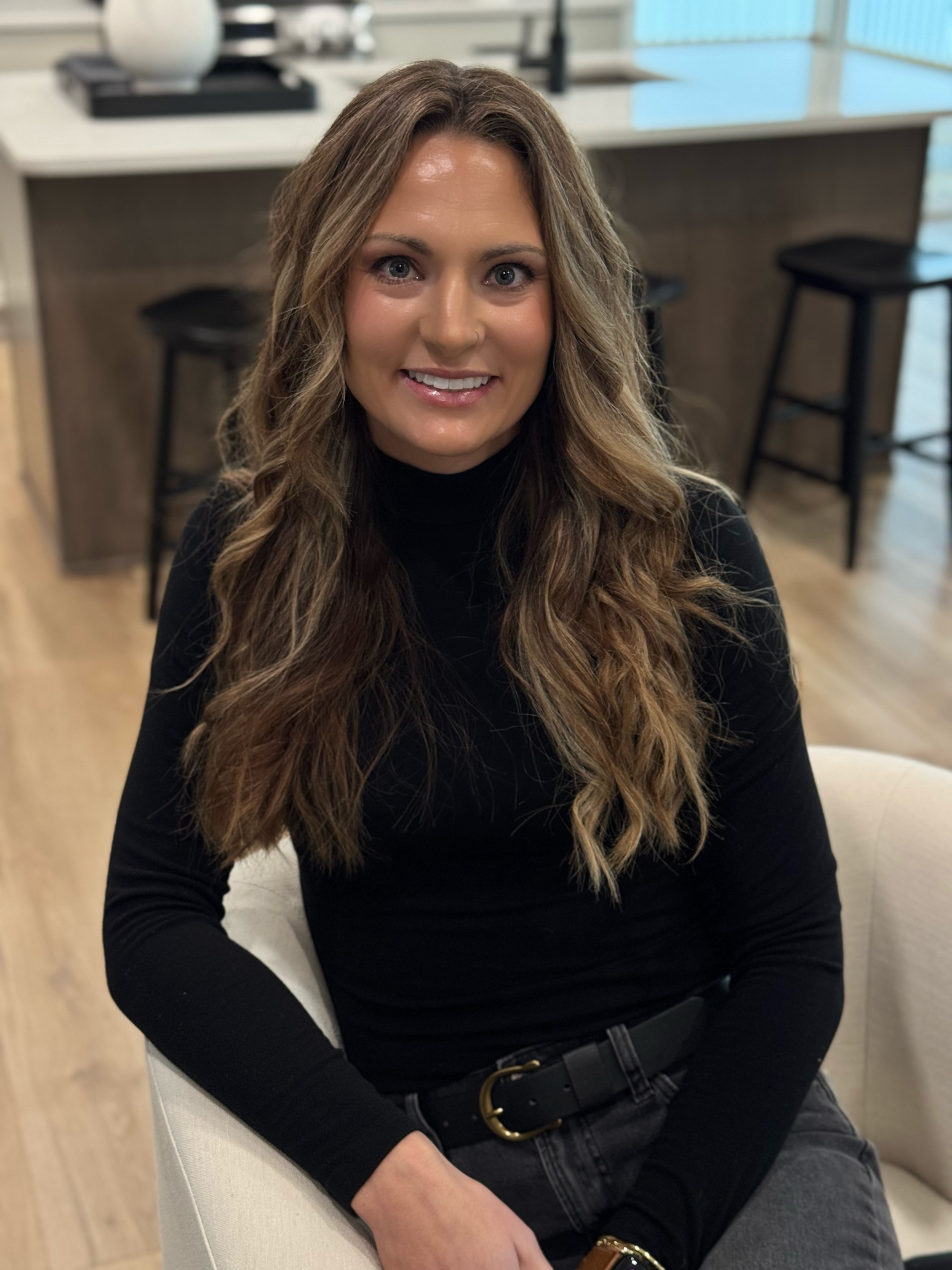 A woman with long wavy brown hair, wearing a black turtleneck, sitting on a white chair in a modern kitchen or dining area.