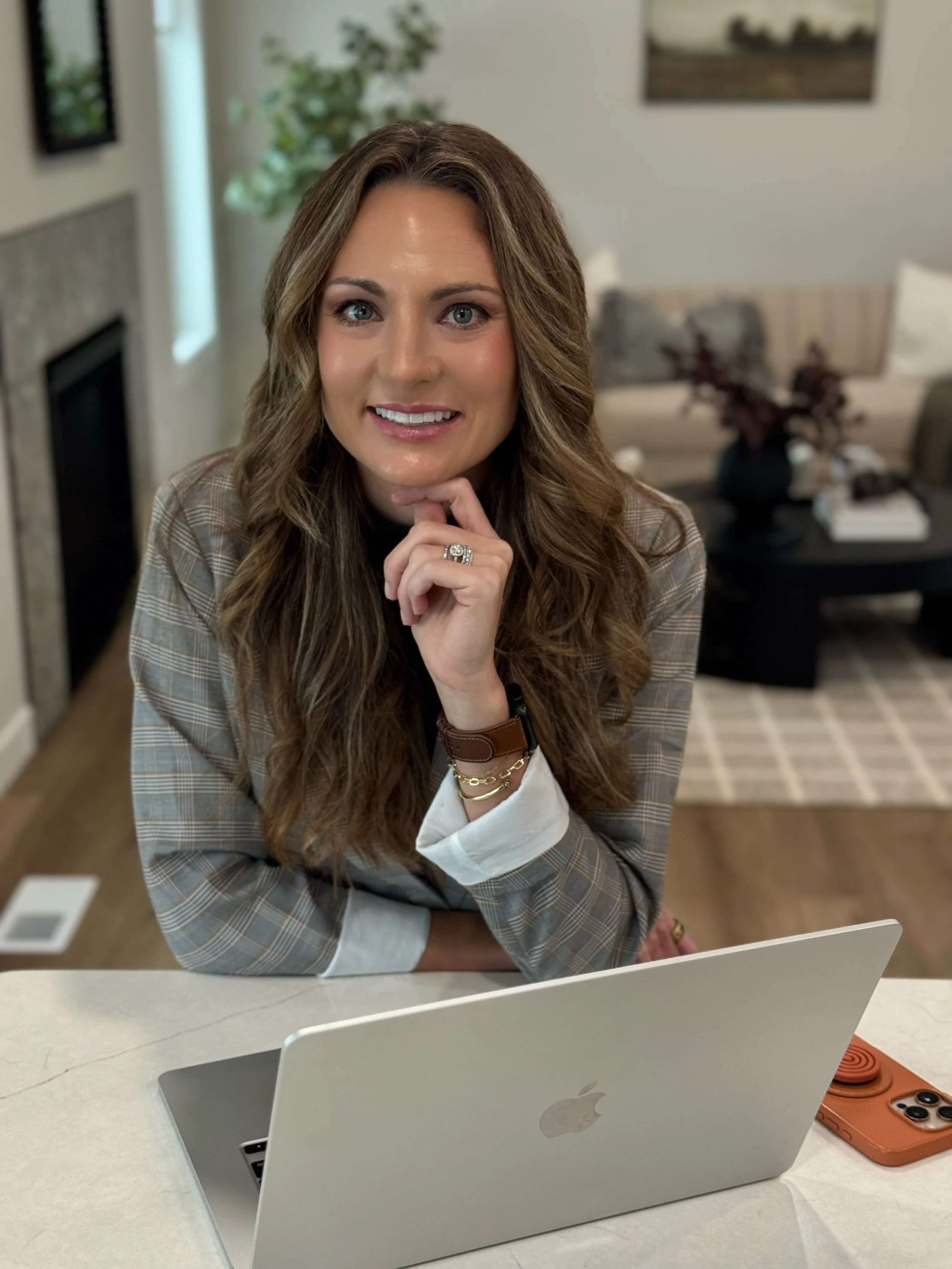 A woman with long wavy brown hair smiling at the camera, sitting at a kitchen counter with a silver MacBook laptop in front of her, wearing a plaid blazer and jewelry, in a cozy living room.