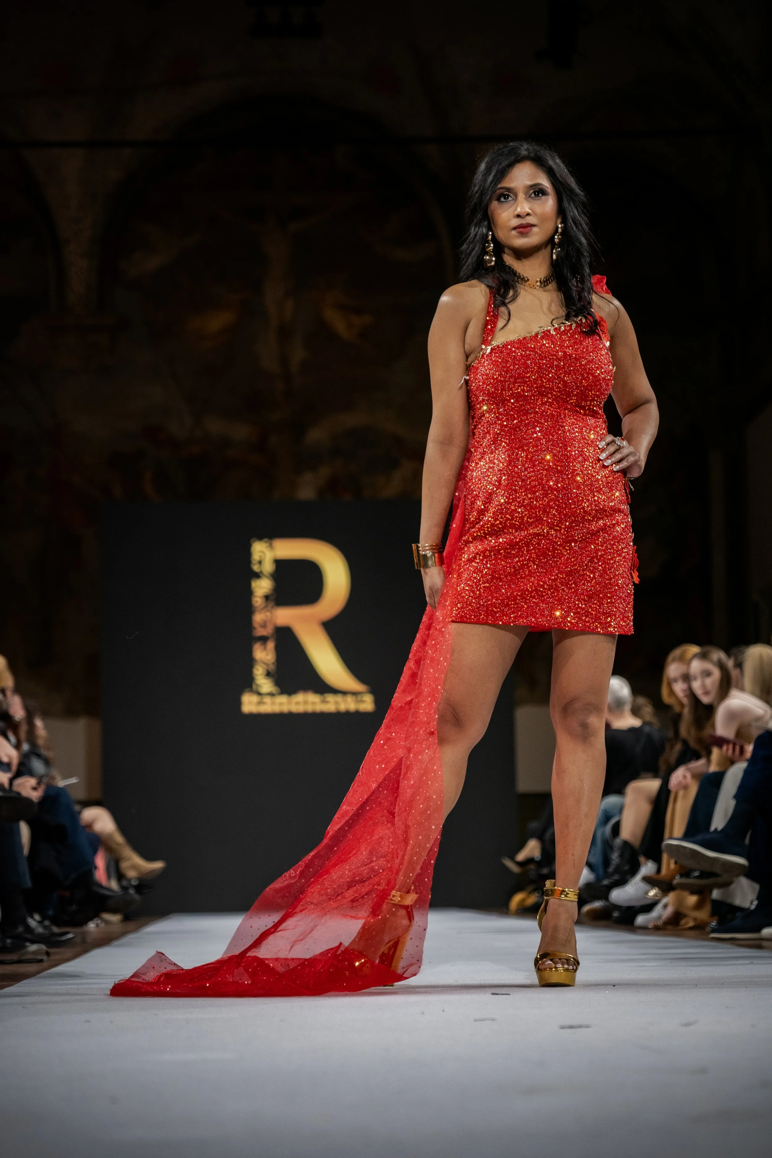 A woman walking on a fashion runway wearing a red, sparkling, asymmetrical dress with a sheer train, gold heels, and jewelry, with an audience watching on either side.